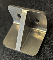 A metal component sample from KT Machining. It is a simple, small bracket with two holes and a triangle dividing the bracket in half.