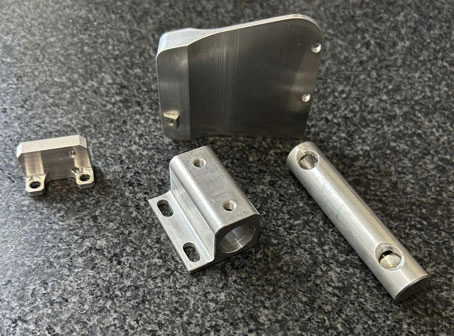 Various small metal components to show the precision that KT Machining can accomplish. A cylinder rod, a flat panel with a taper, a rectangle with a flat lip and a hole for a rod, and a small piece with holes.
