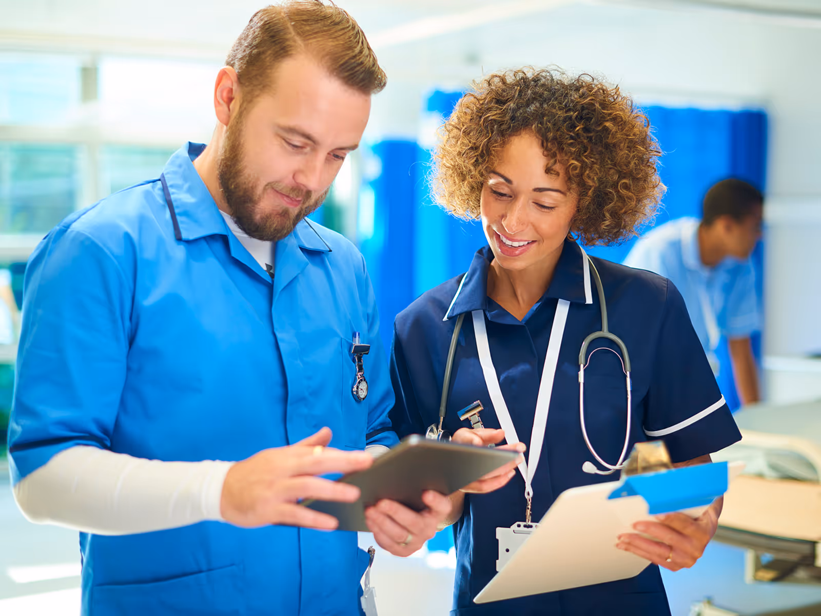 Two healthcare professionals in blue scrubs review a tablet together in a bright clinical setting, with blue curtains and a blurred staff member in the background, conveying teamwork and communication.