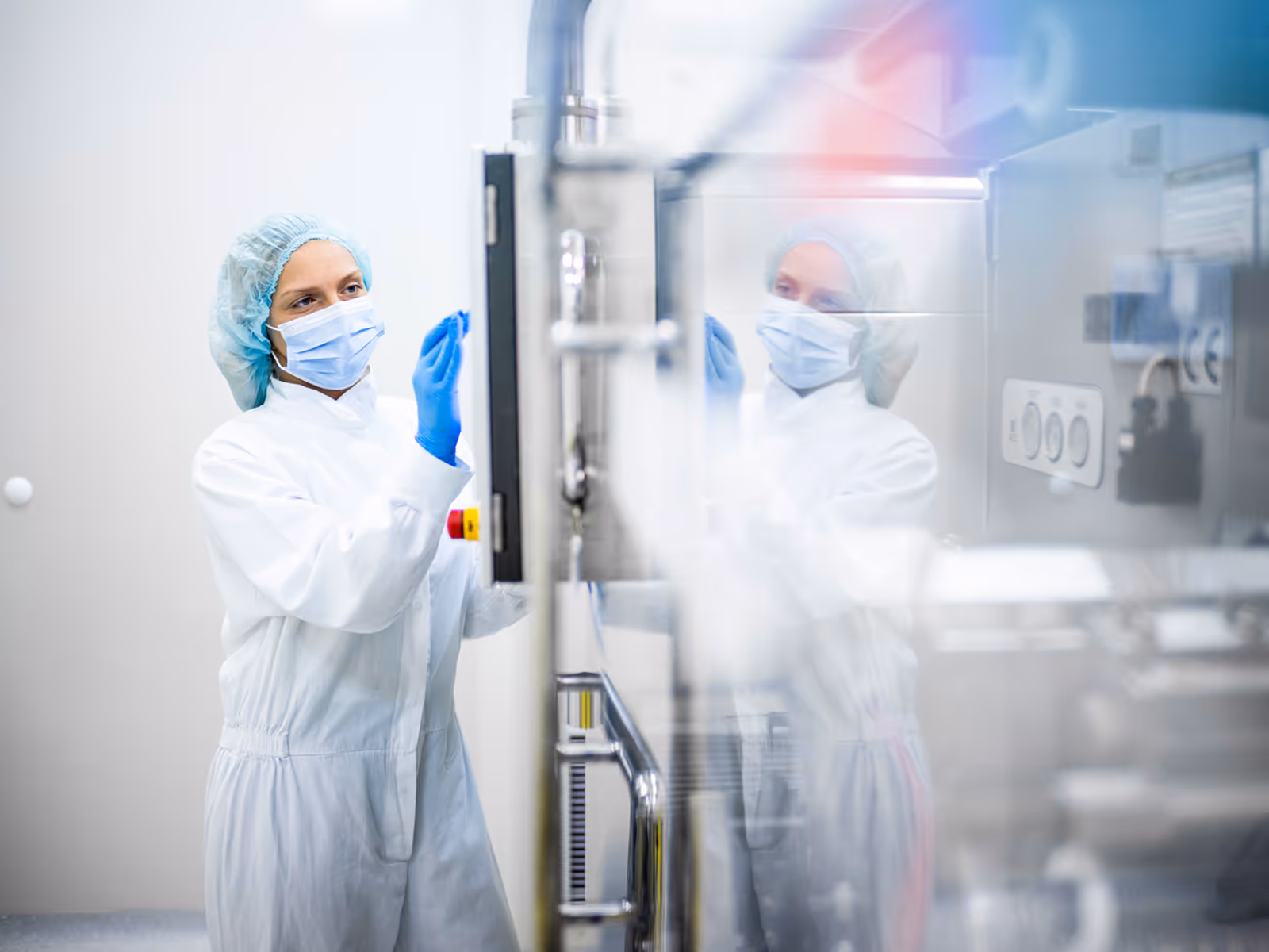 A cleanroom technician in a white coat, blue mask, gloves, and hairnet adjusts laboratory equipment, with controls and indicators visible on a sterile, light-colored background.