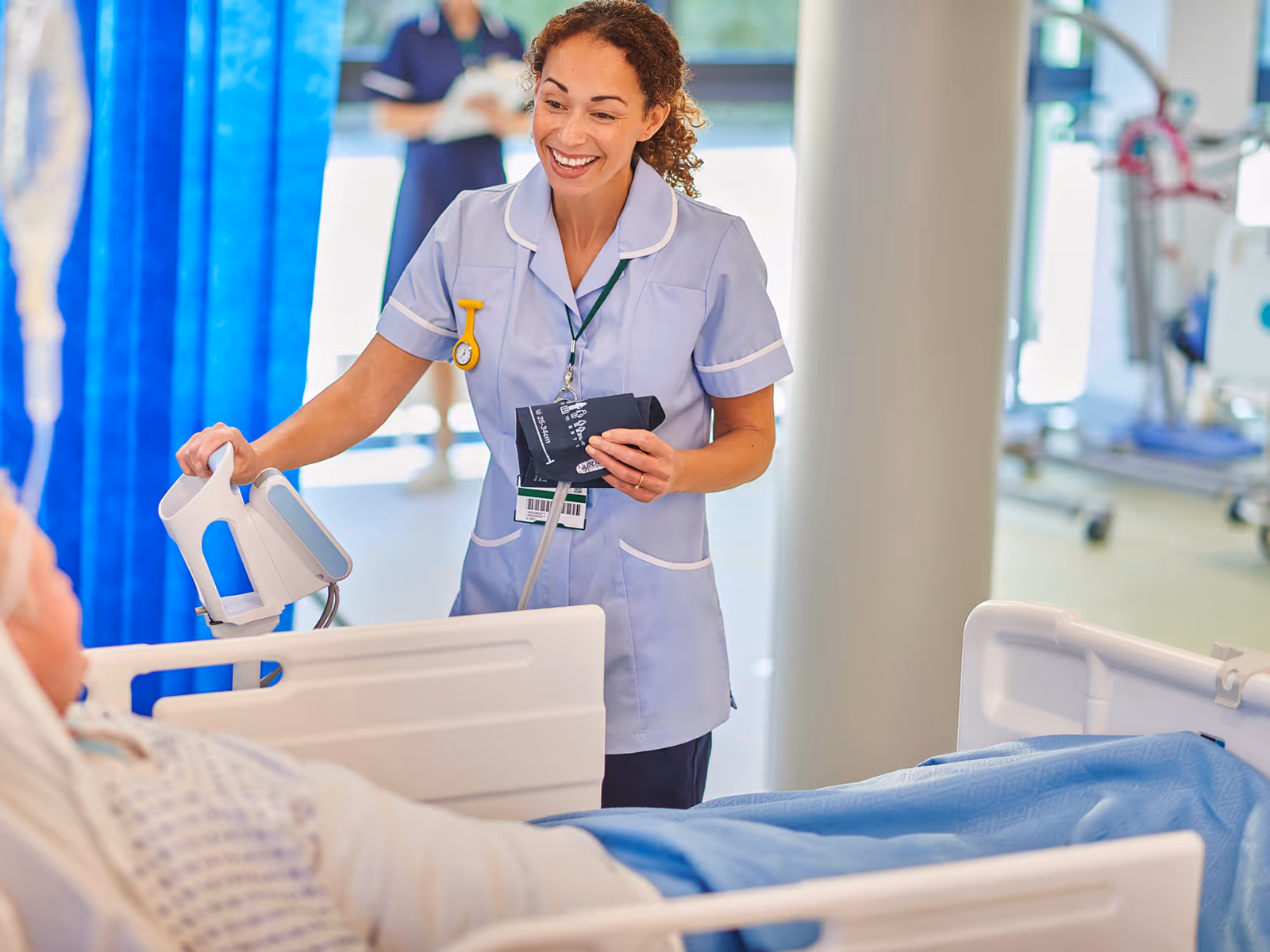 A friendly nurse in a light blue uniform smiles while checking a patient’s bedside monitor in a bright hospital ward with blue curtains and staff in the background.