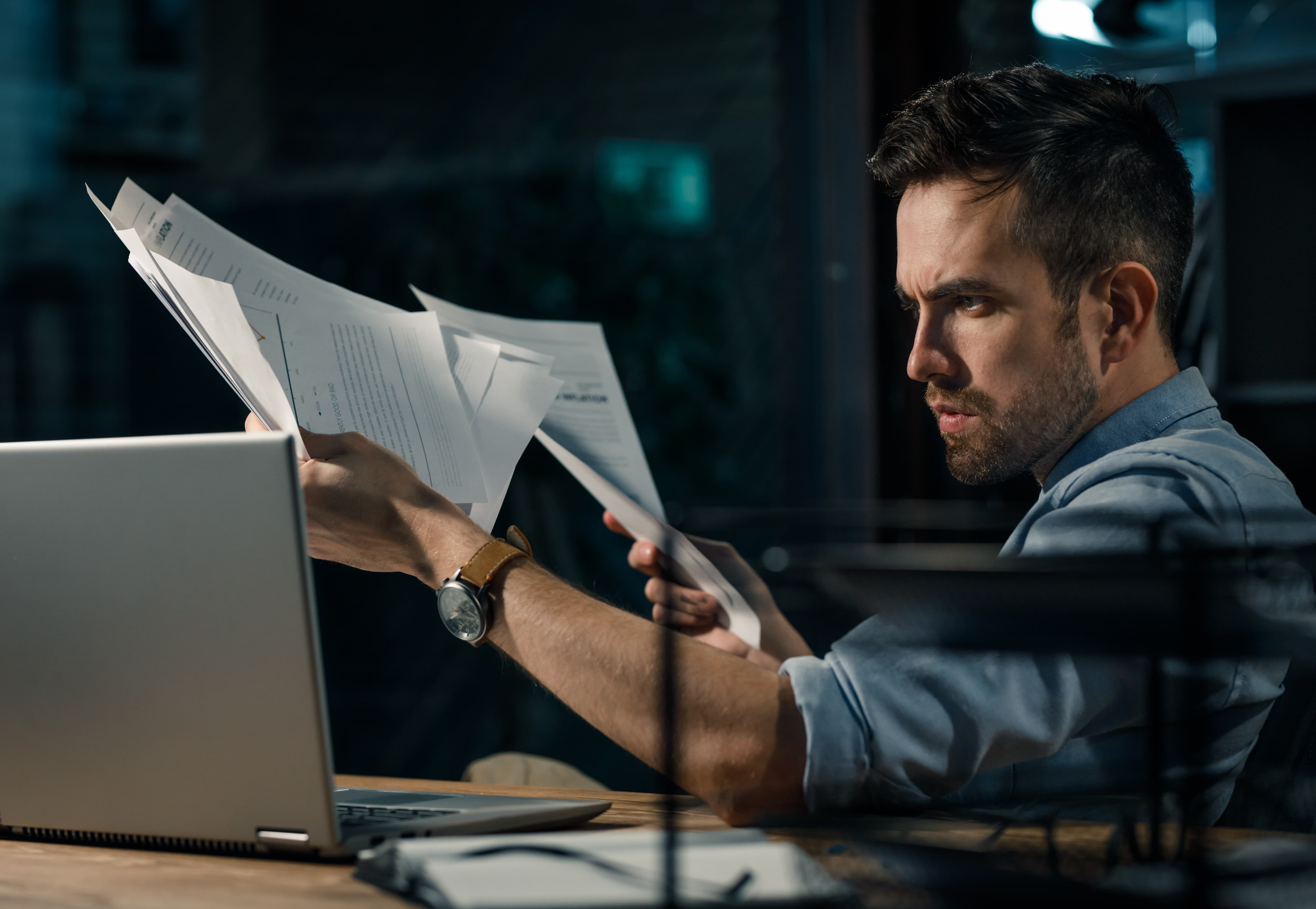 Young man in shirt holding pile of papers and documents while sitting at table and looking at laptop seriously.