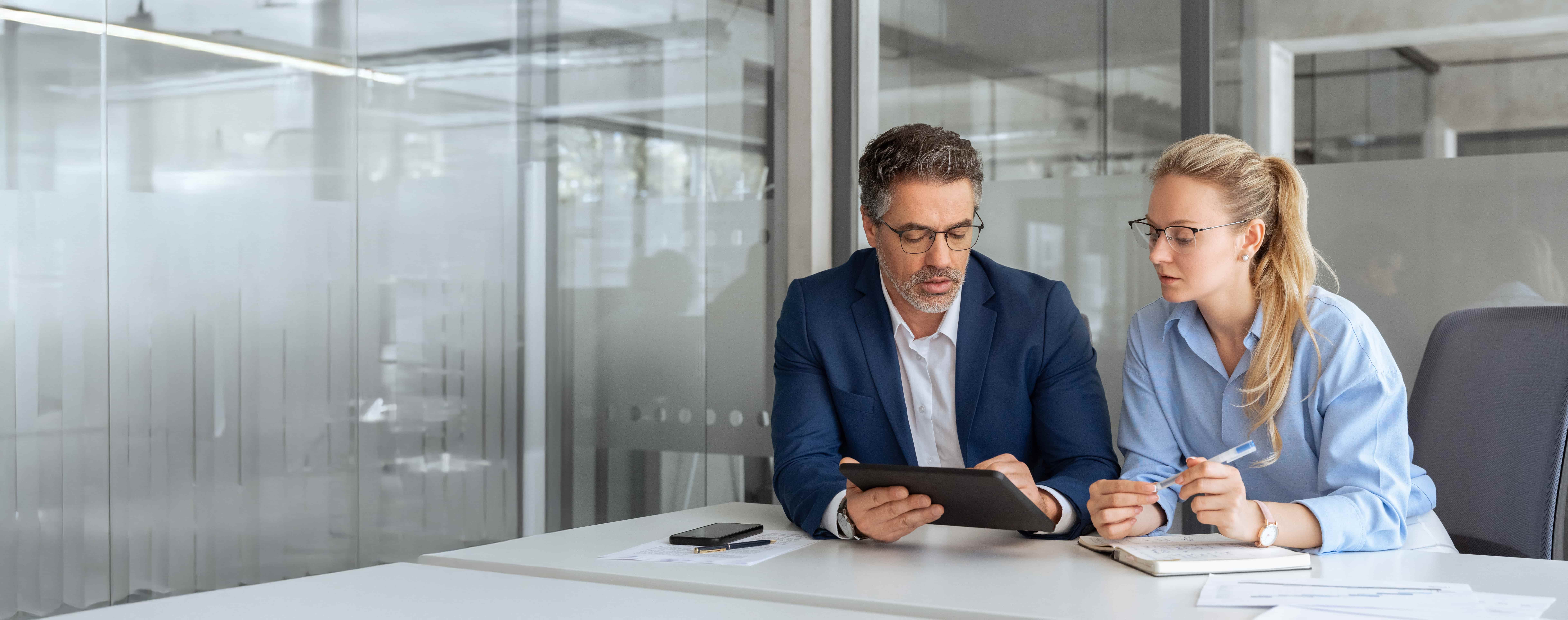 Two professional business people working together. Partners mature man and woman discussing project in office. Team leader explain trainee working process using tablet pc computer. 