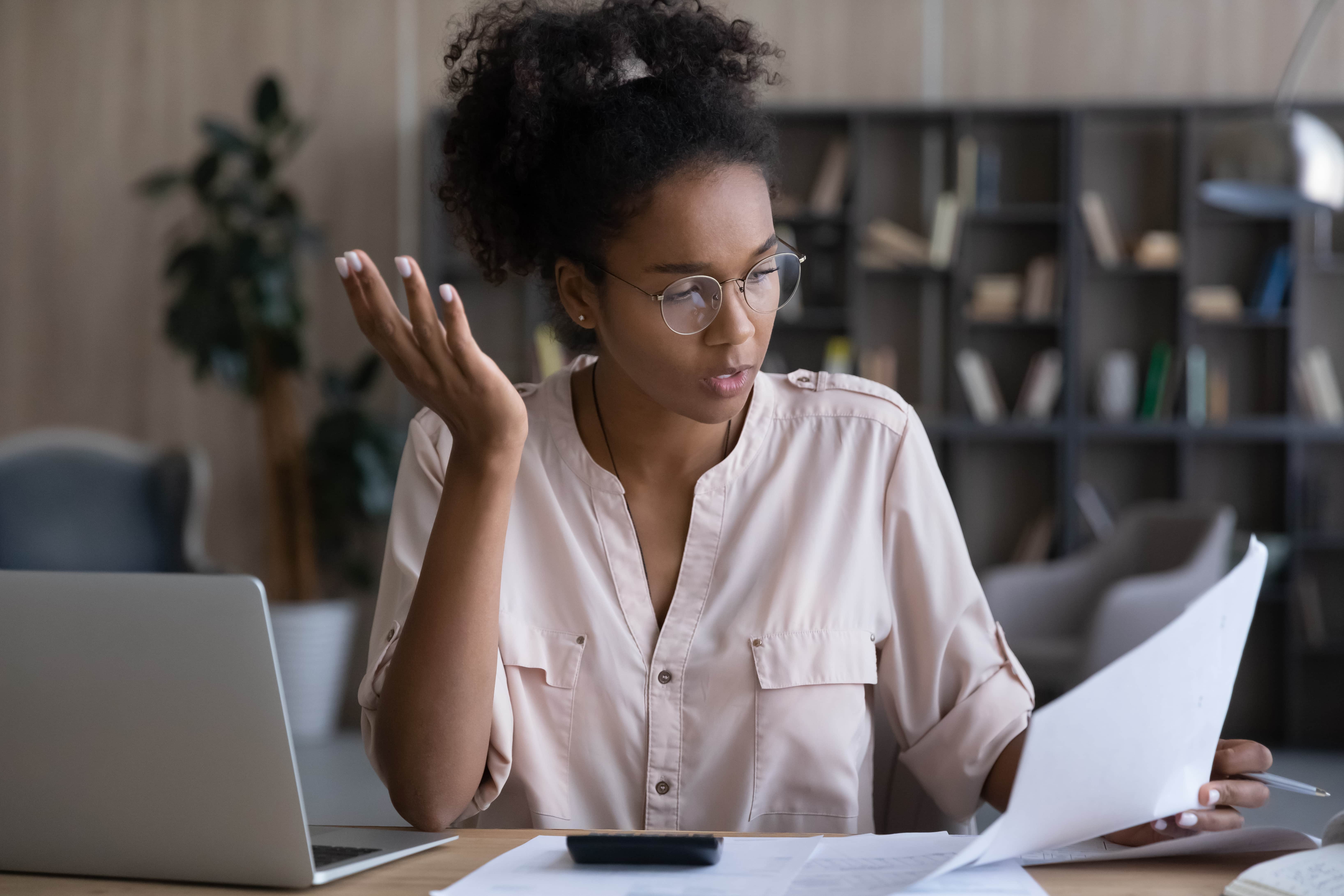 Serious Black woman reading and checking wrong financial papers, bills for paying with mistakes. Confused puzzled accountant reviewing tax documents for payment at workplace with laptop