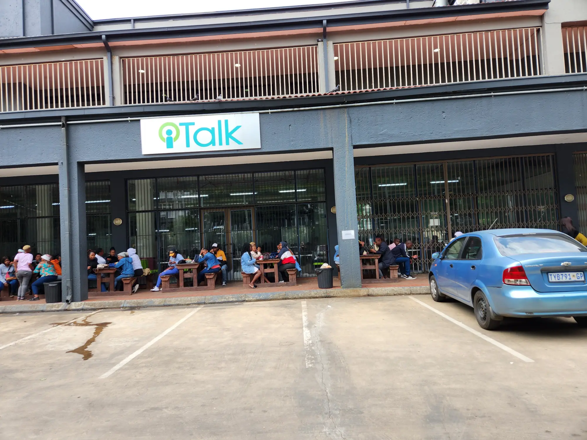 People sitting at outdoor wooden tables under a building sign reading iTalk, with a blue car parked nearby.