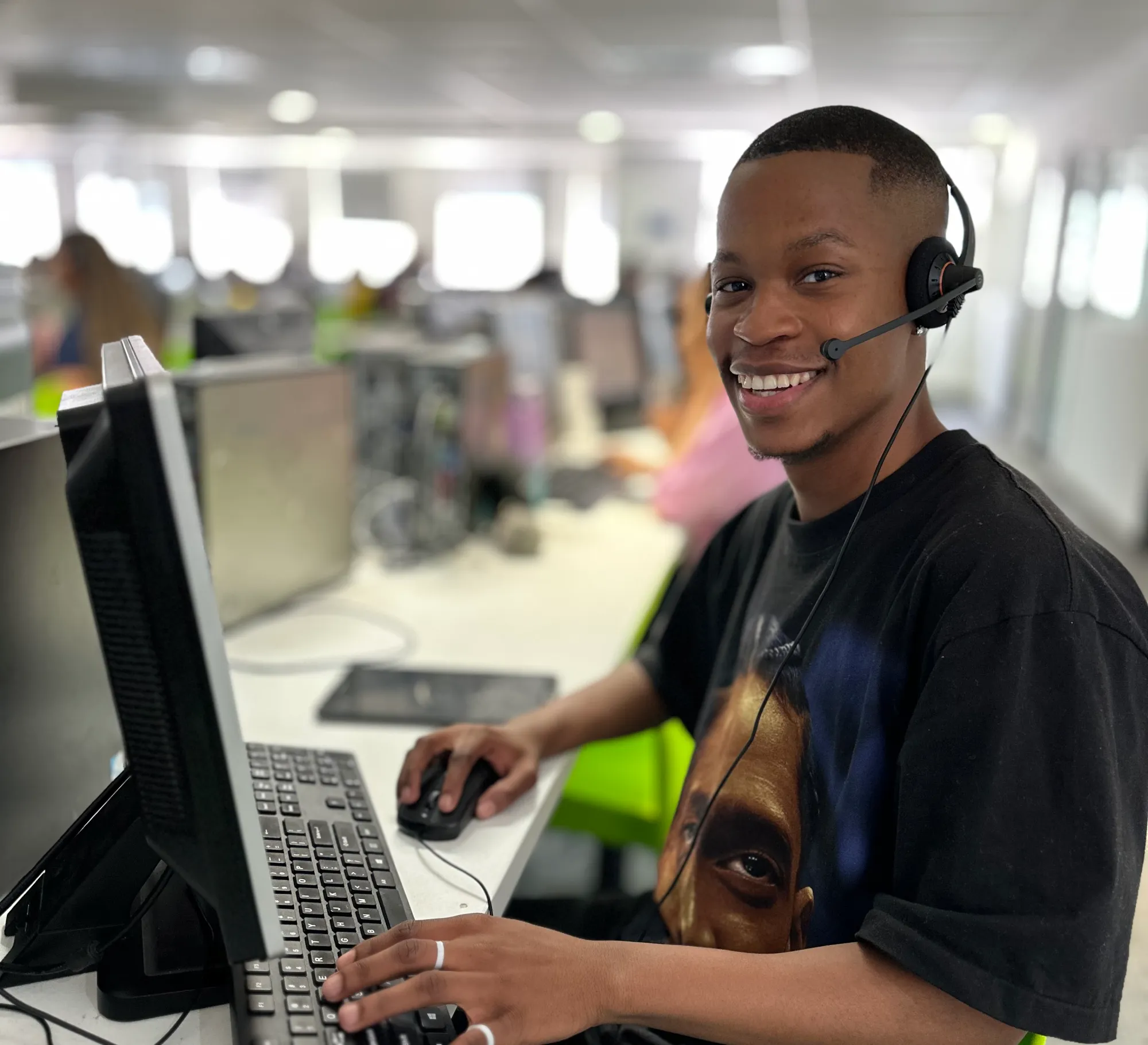 Smiling man wearing headset uses computer mouse and keyboard in a bright office setting.