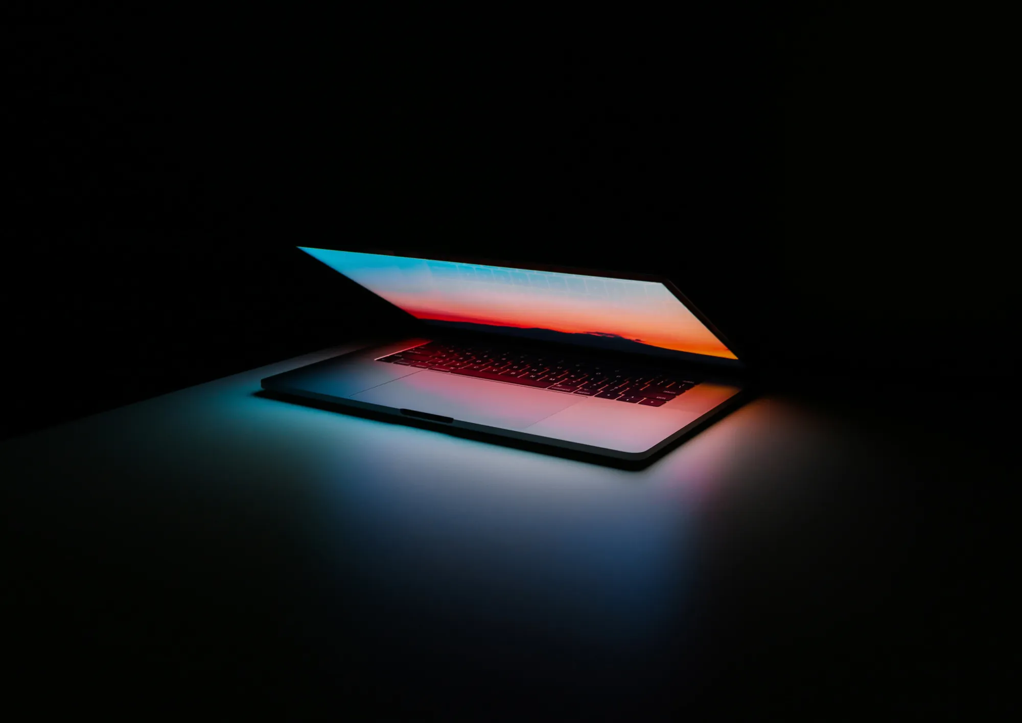 Partially open laptop in dark room showing colorful sunset or sunrise on screen and glowing keyboard.
