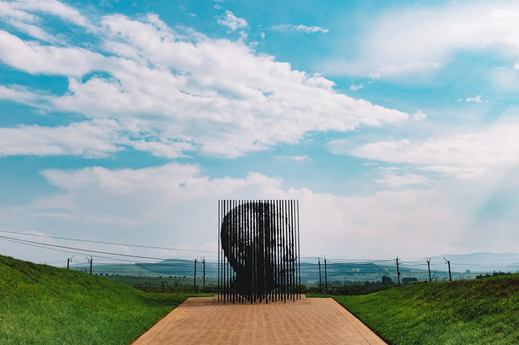 Sculpture of a head made from vertical black rods on a walkway with grass and blue sky background.