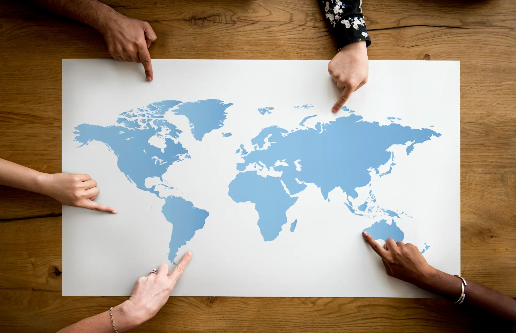 Four diverse hands pointing at different continents on a blue world map on a wooden table.