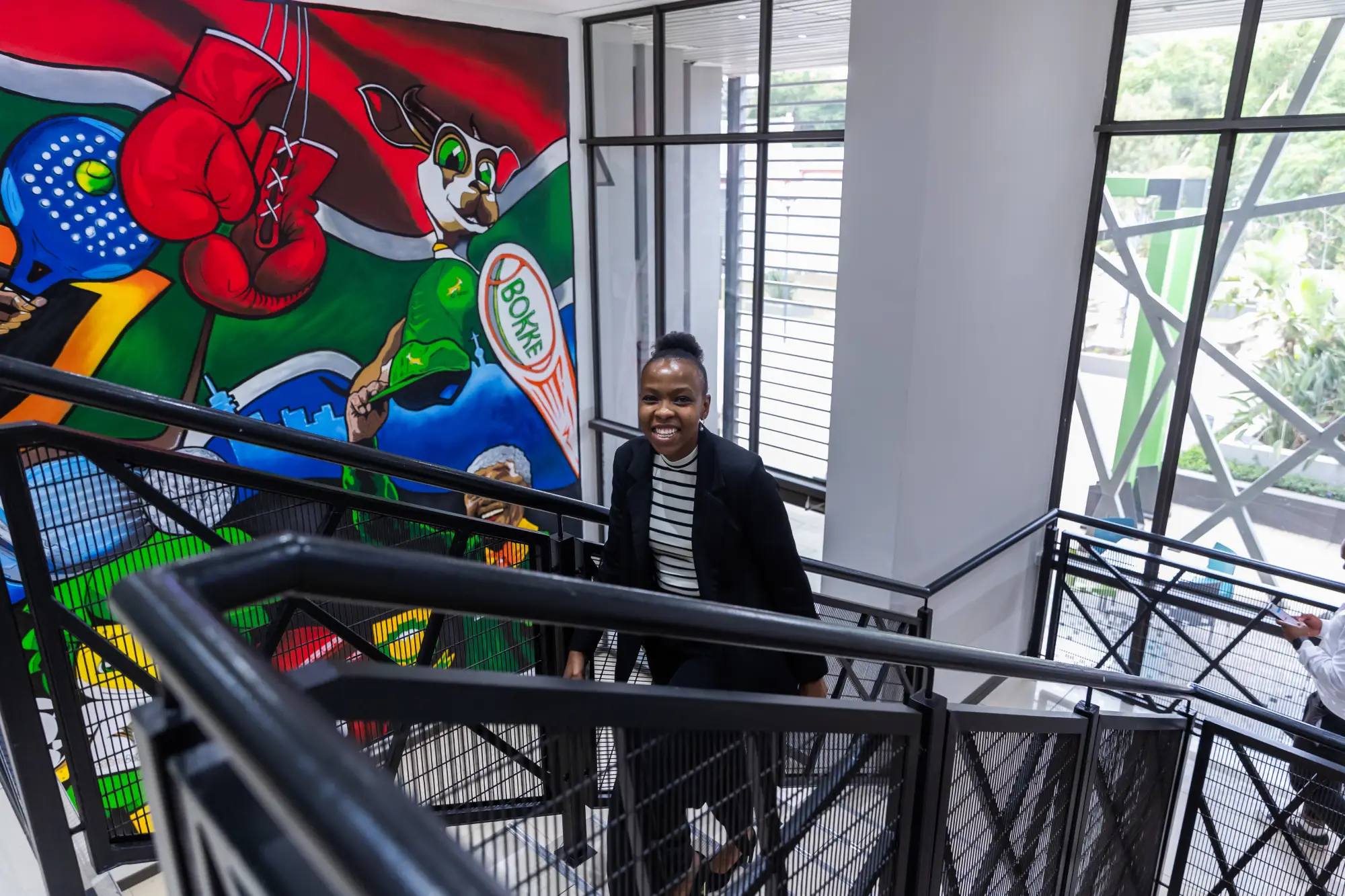 Smiling woman in black blazer ascending staircase with colorful sports mural on wall behind her.