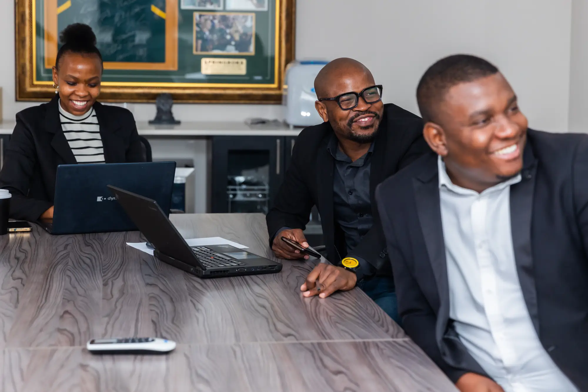 Three smiling professionals in business attire with laptops around a large wooden conference table.