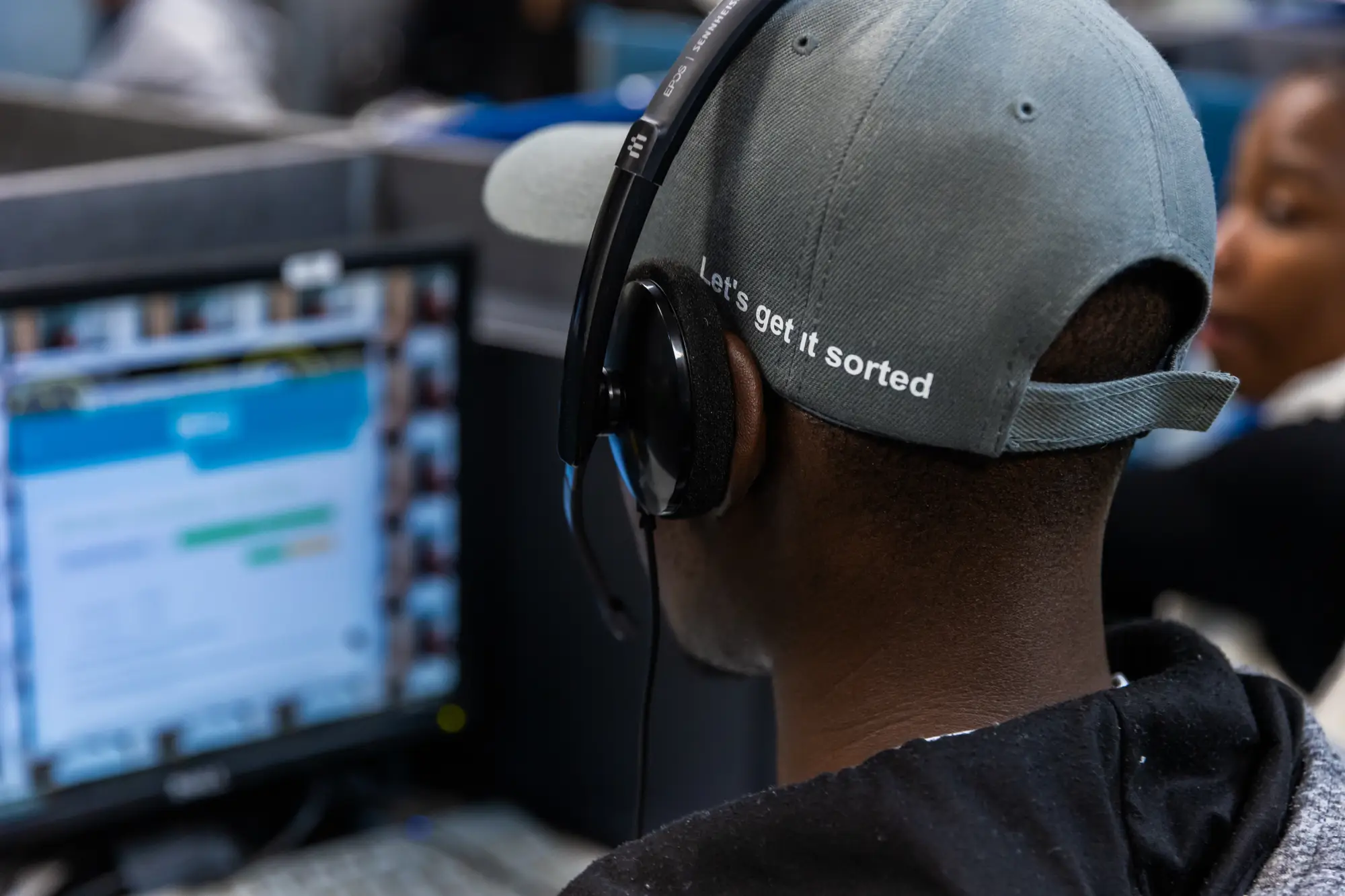 Person wearing gray cap with 'Let's get it sorted' and headset, looking at a blurred computer screen.