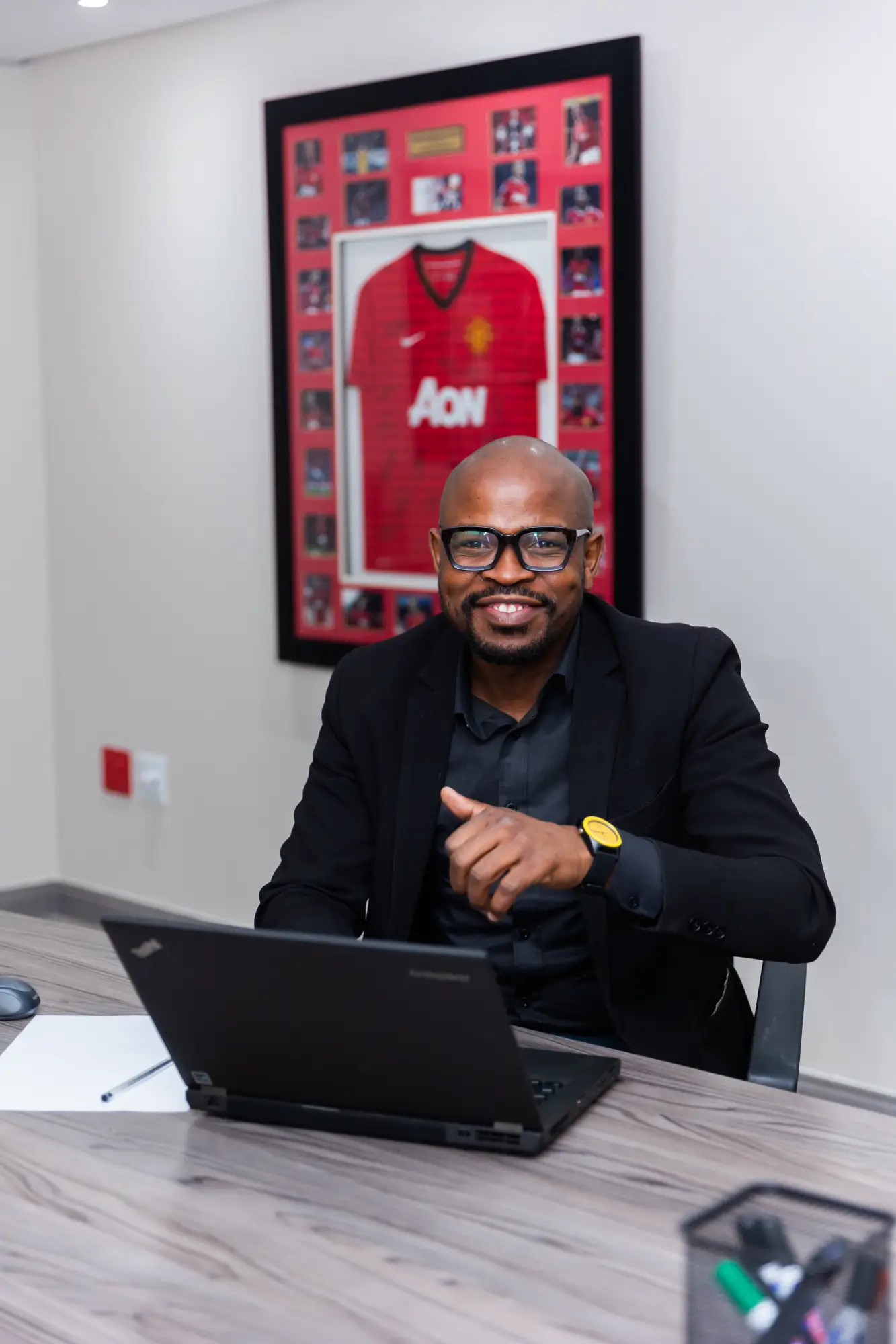 Smiling man in black blazer and glasses sitting at desk with laptop, framed soccer jersey behind him.