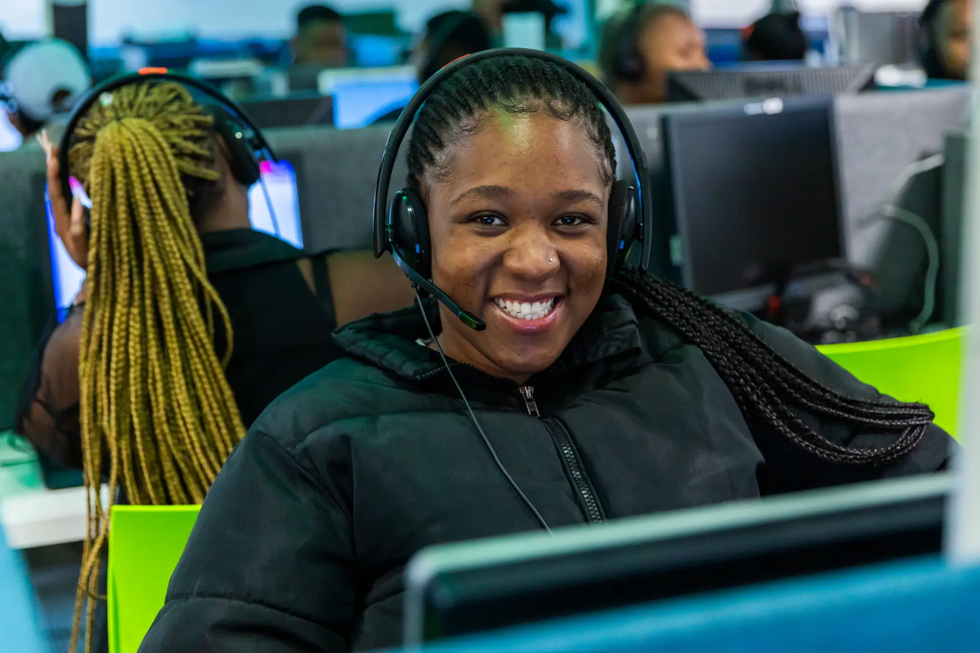 Smiling customer service agent wearing a headset and black jacket at a call center workstation.