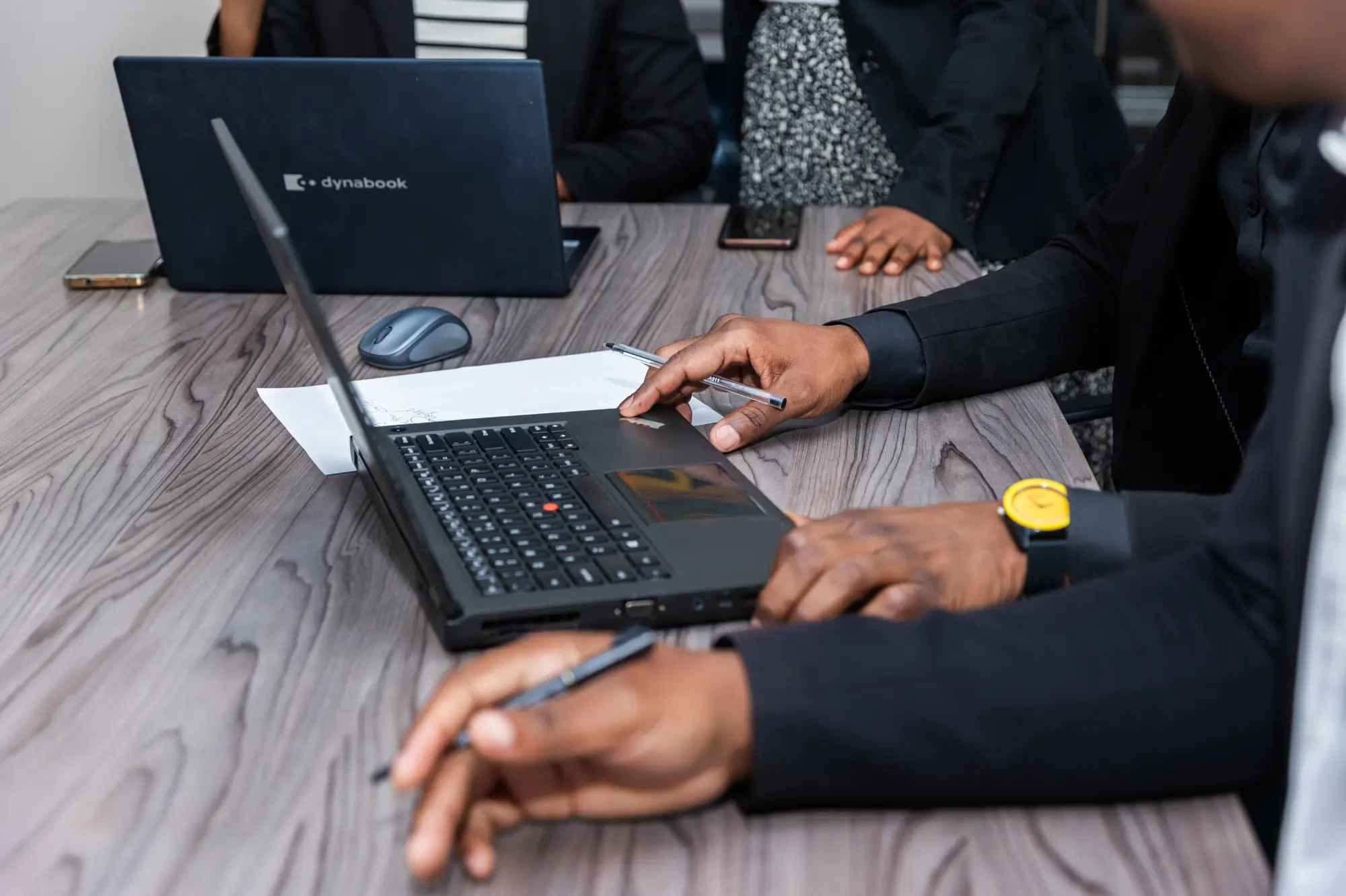 People in business attire using laptops and holding pens around a wooden conference table.
