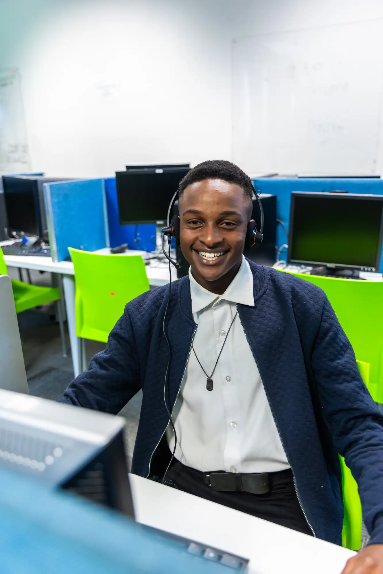 Smiling man wearing headset sitting at a computer in a modern office with green chairs.