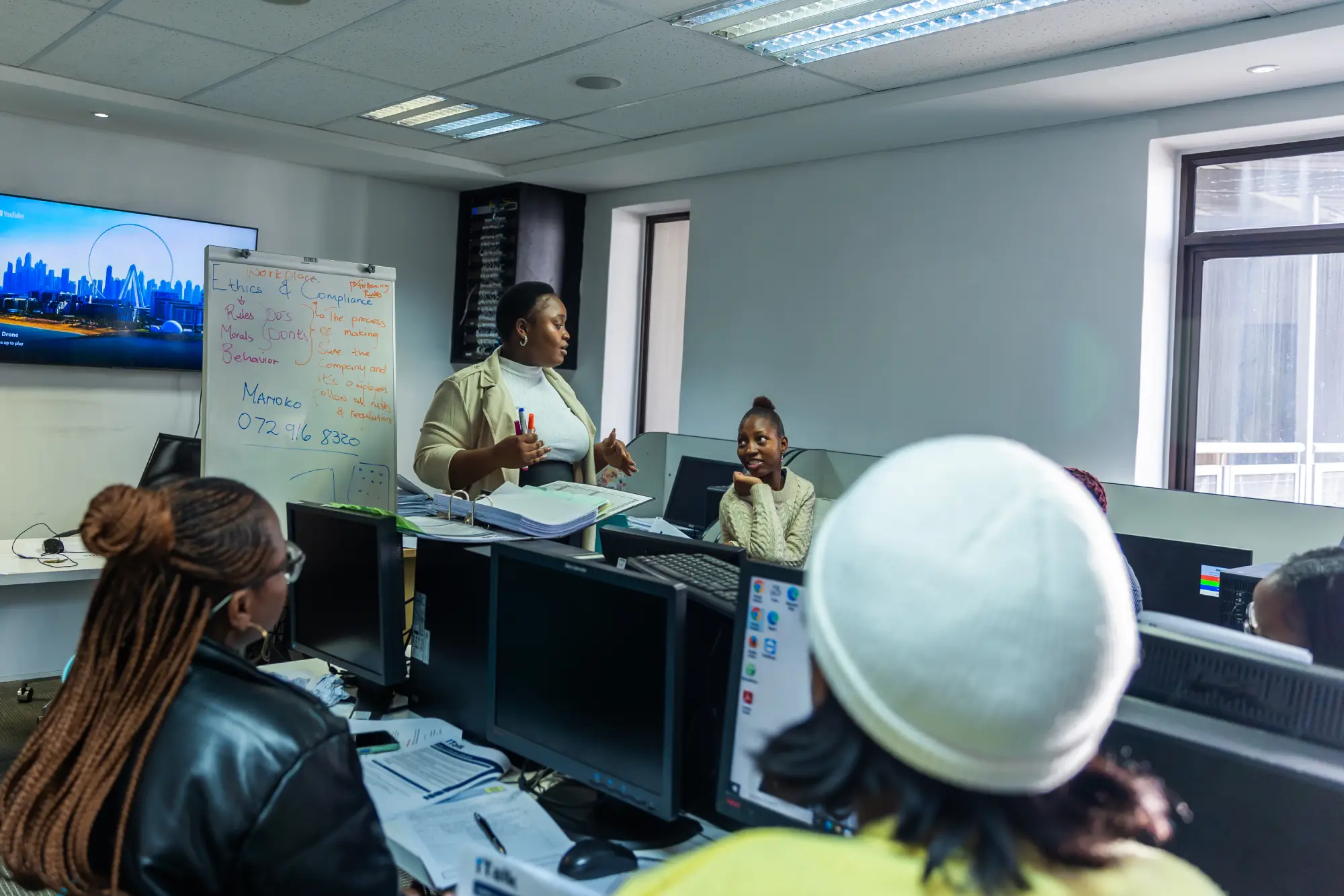 Woman standing explaining to coworkers in an office with computers and whiteboard notes.