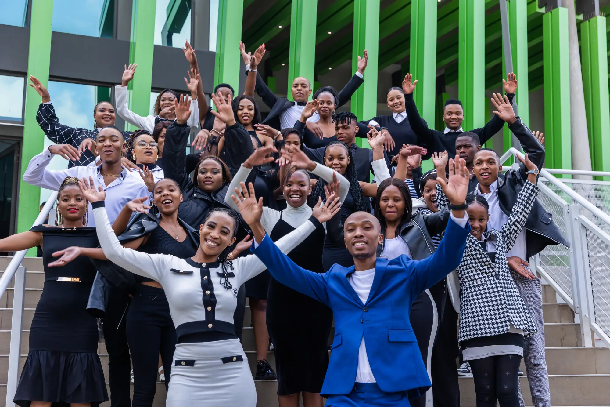 Happy diverse group of adults waving with raised arms outside a green modern building on steps.