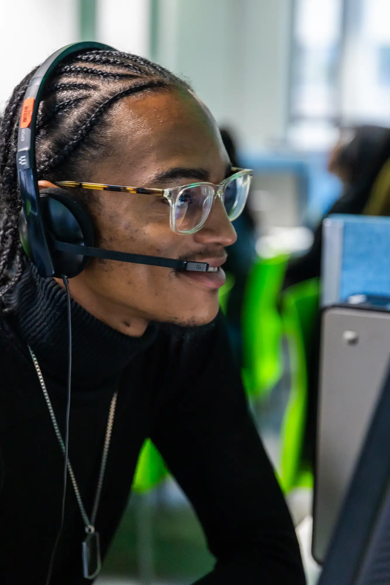Smiling person with braided hair and glasses wearing a headset, working at a computer.