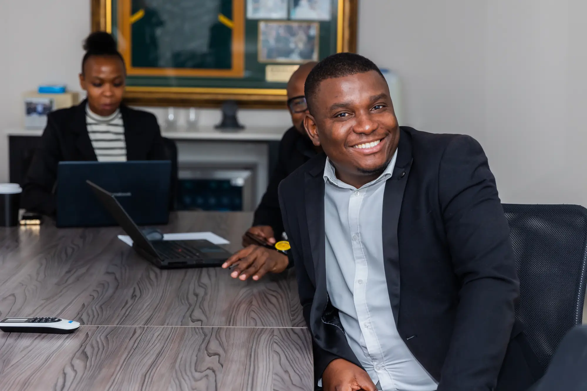 Smiling man in a suit seated at a table with blurred coworkers and laptops in the background.