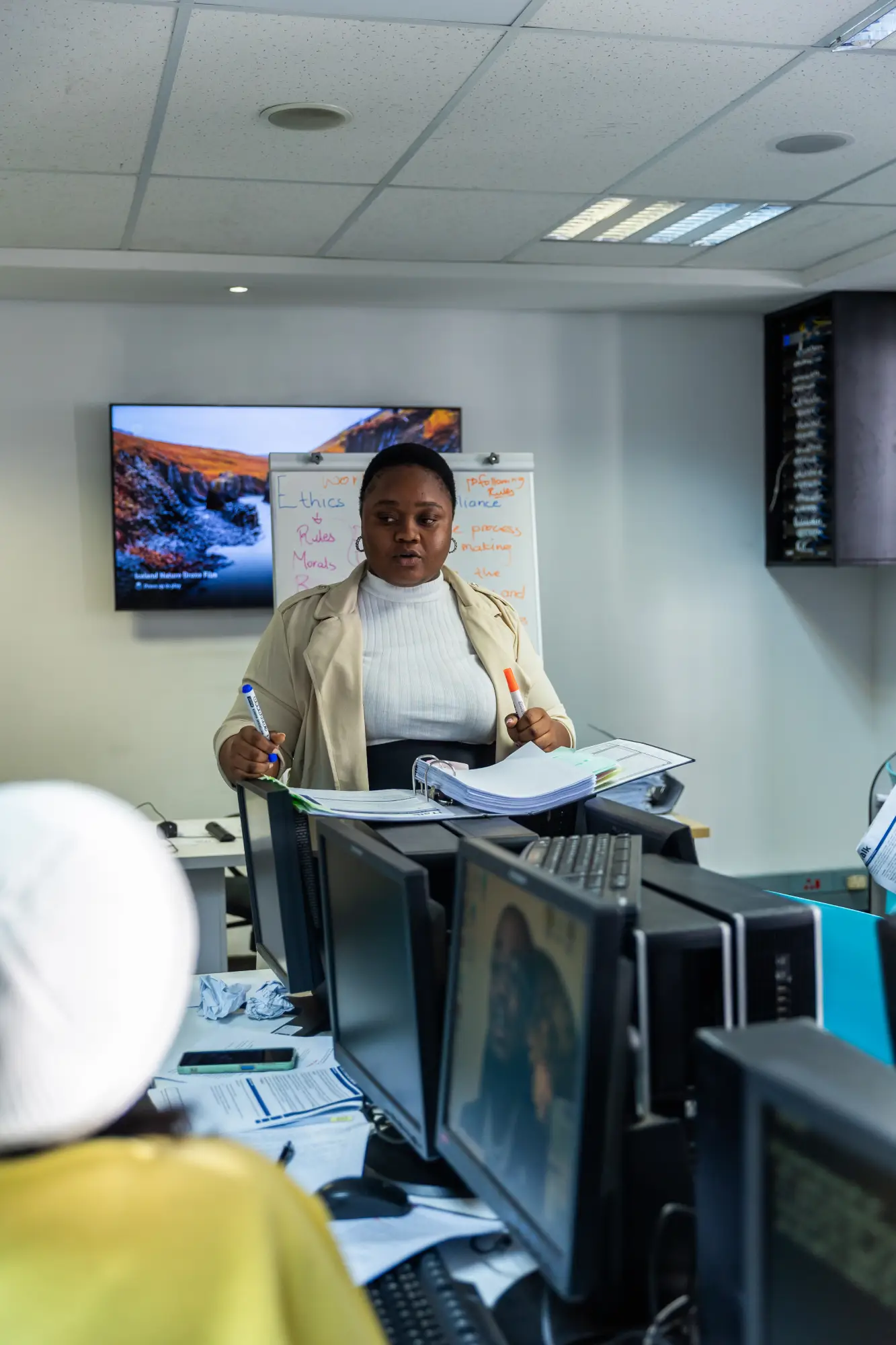 Woman holding markers and papers stands in front of a whiteboard and multiple computer monitors in an office.