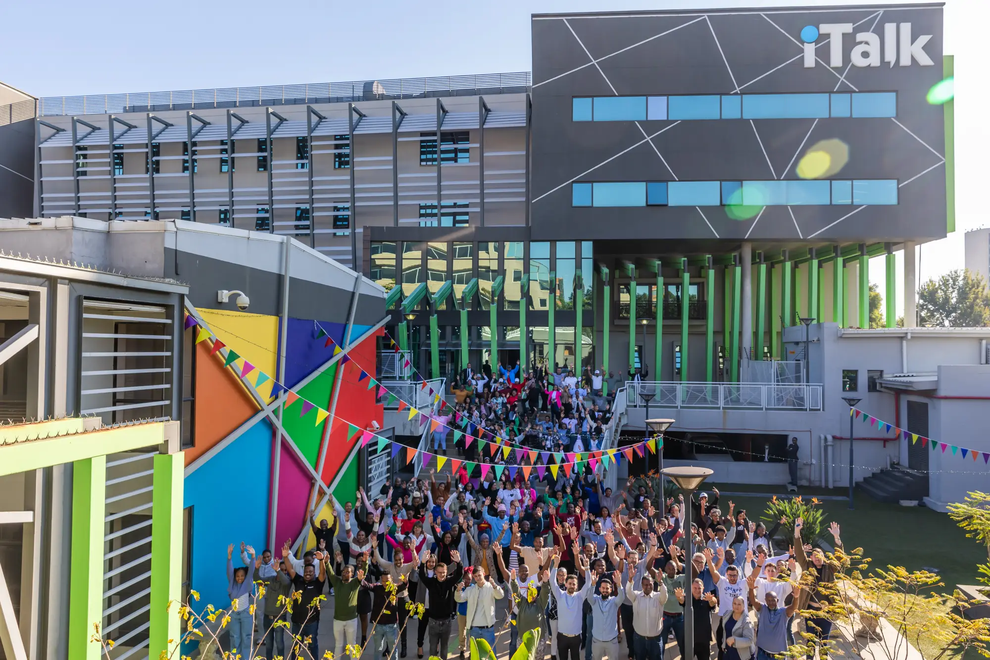 Large diverse group of people raising hands outside colorful modern building with iTalk logo and flags.