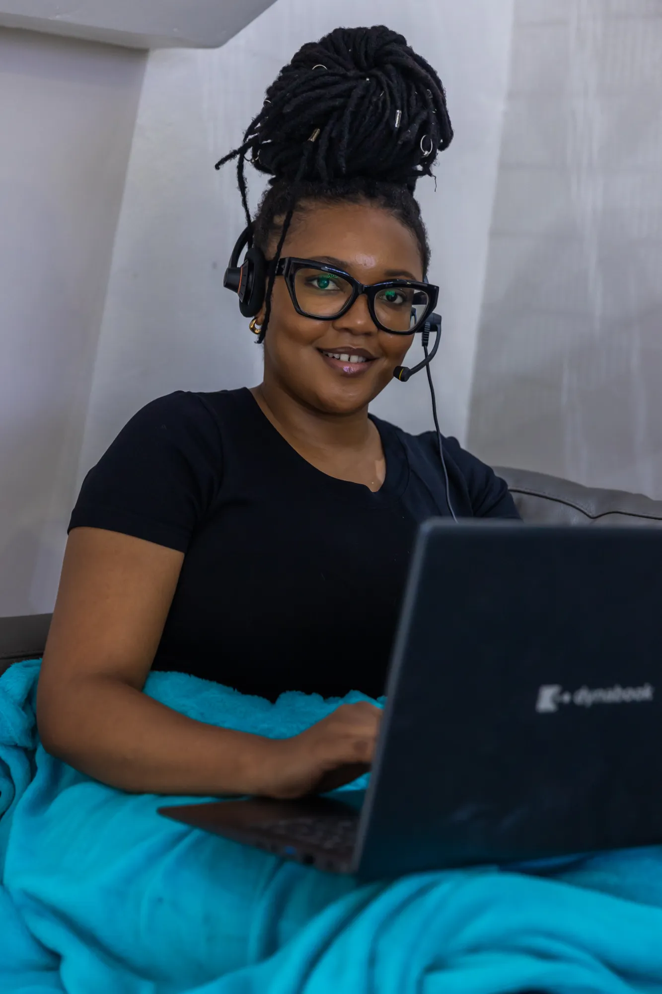 Smiling woman with glasses and headset working on a laptop while wrapped in a blue blanket.