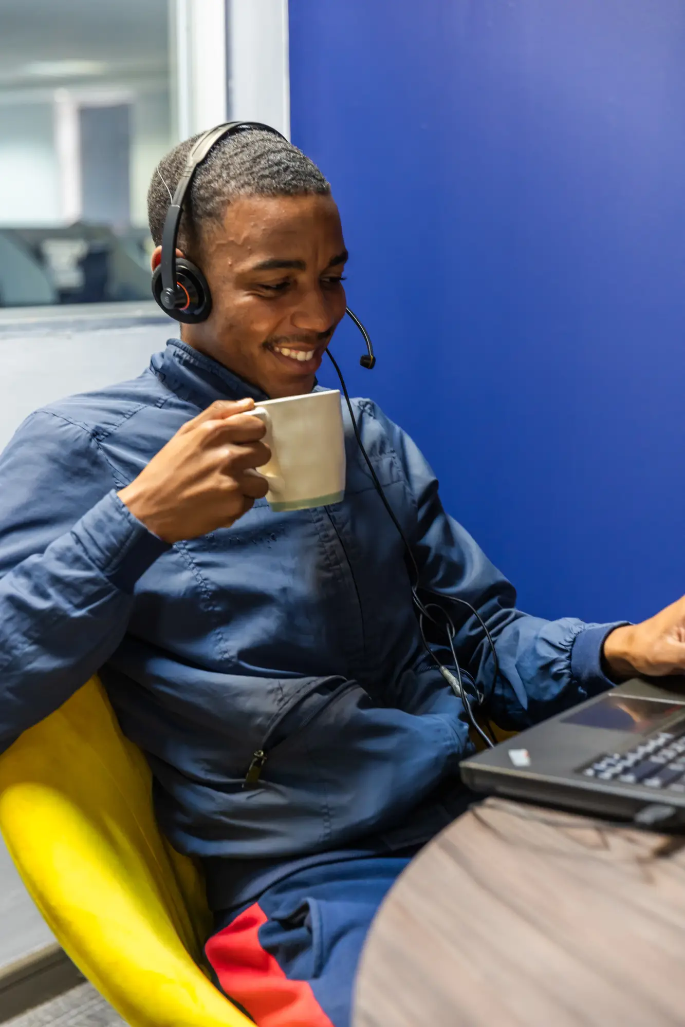 Man wearing headset smiles while holding a mug and using laptop at a table.