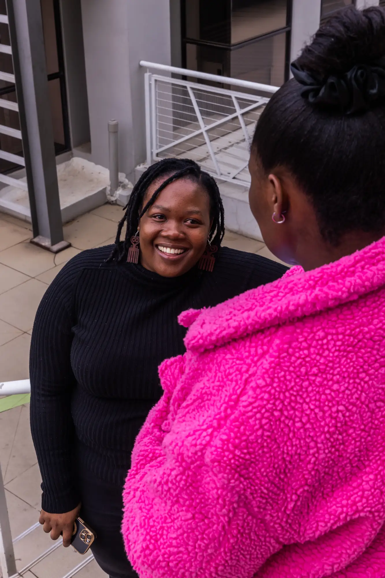 Smiling woman in black sweater holding a phone talks to someone in bright pink coat.