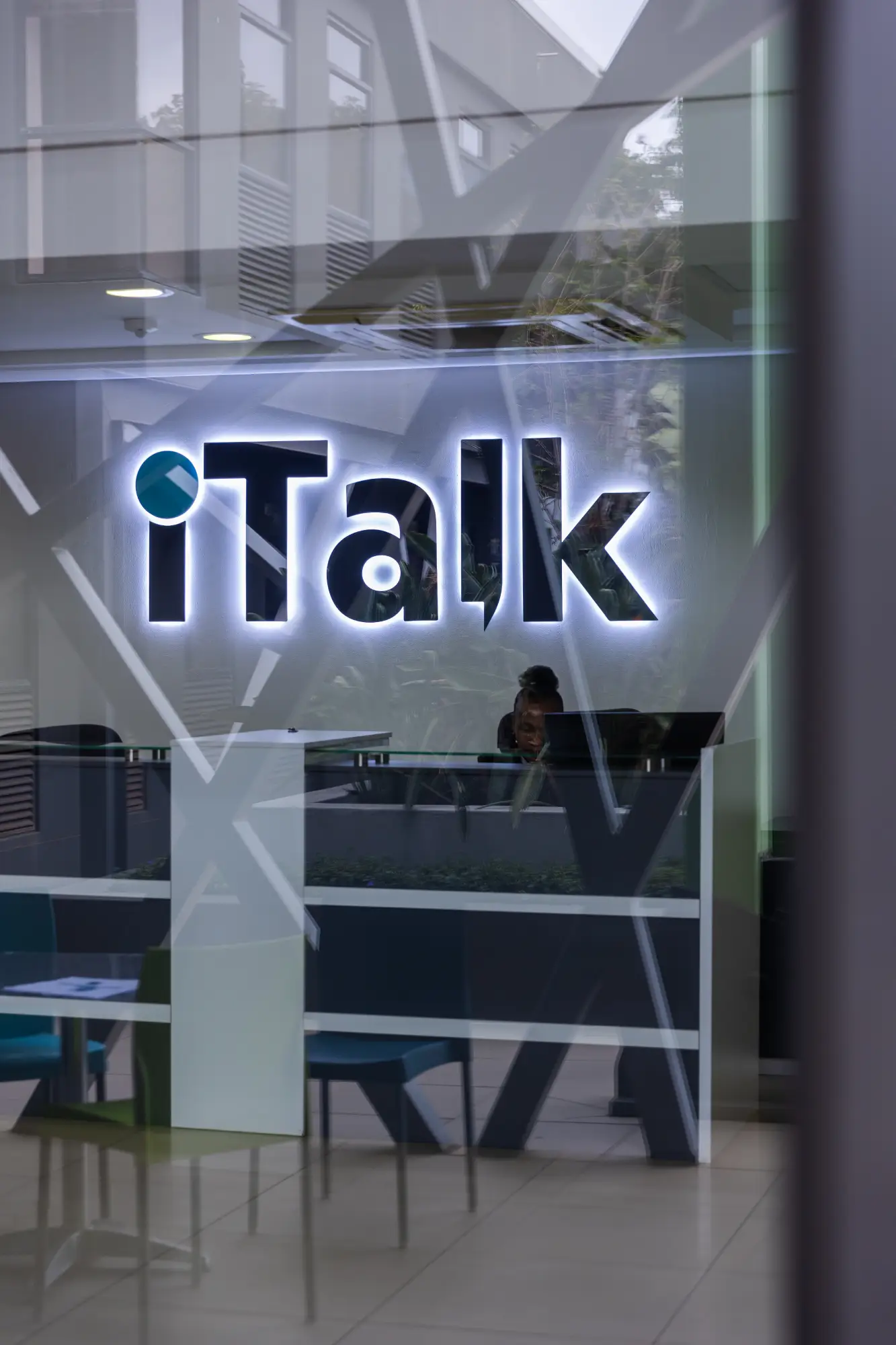 Reception desk inside office with illuminated iTalk logo on the wall and a person seated behind it.