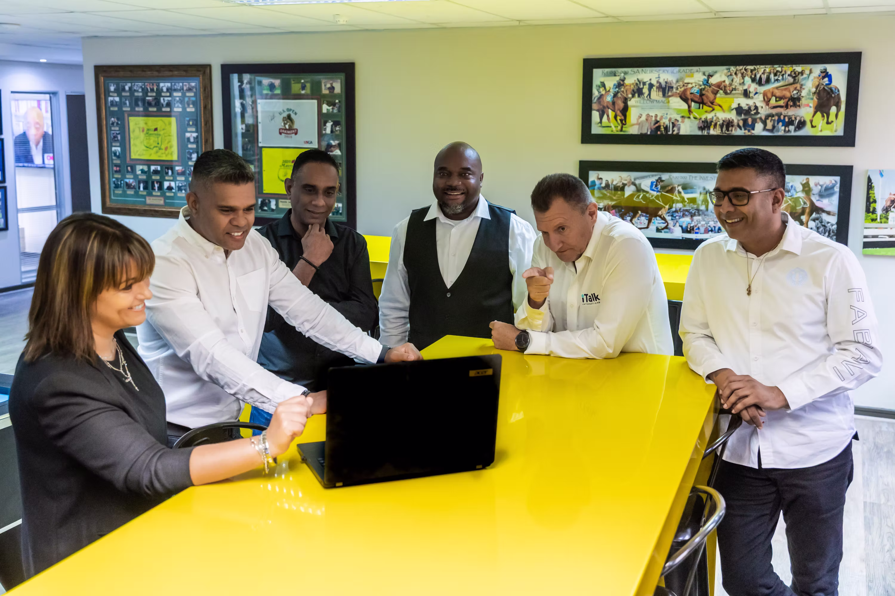 Six coworkers smiling and engaged around a yellow table looking at a laptop in an office.