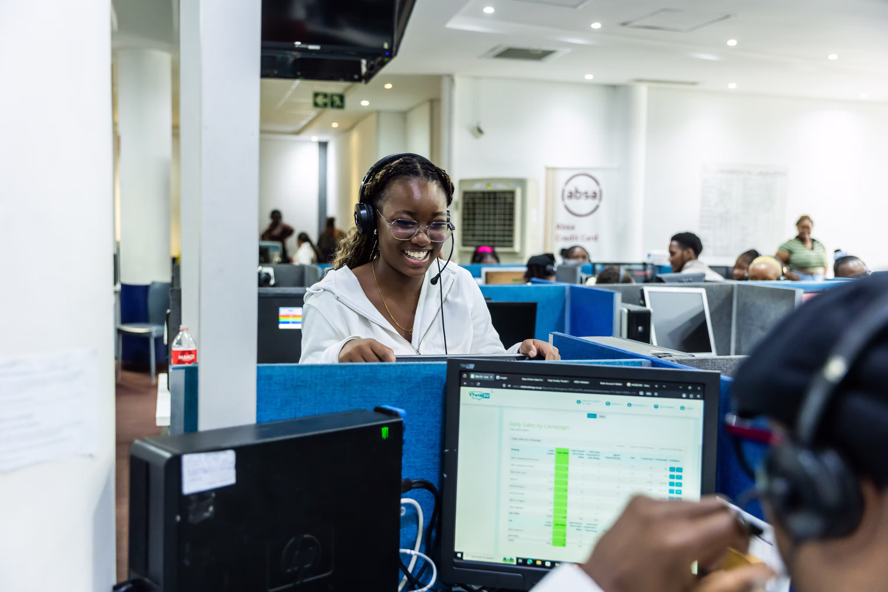 Smiling woman with headset and glasses working at a computer in a busy call center office.