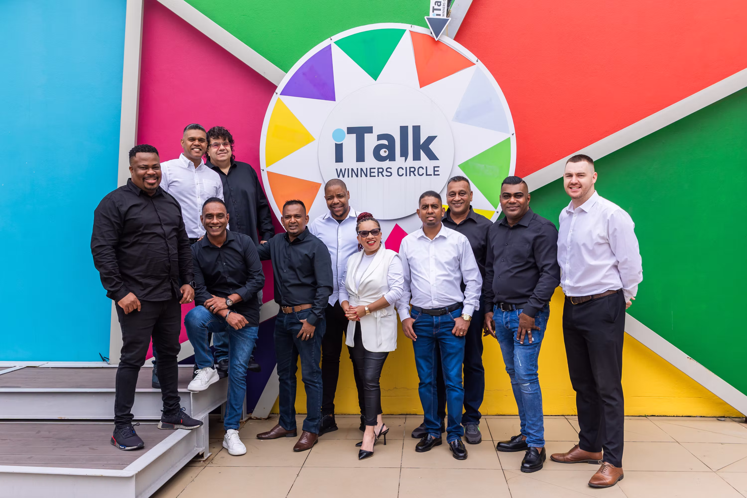 Group of people posing in front of colorful wall with iTalk Winners Circle logo wheel.