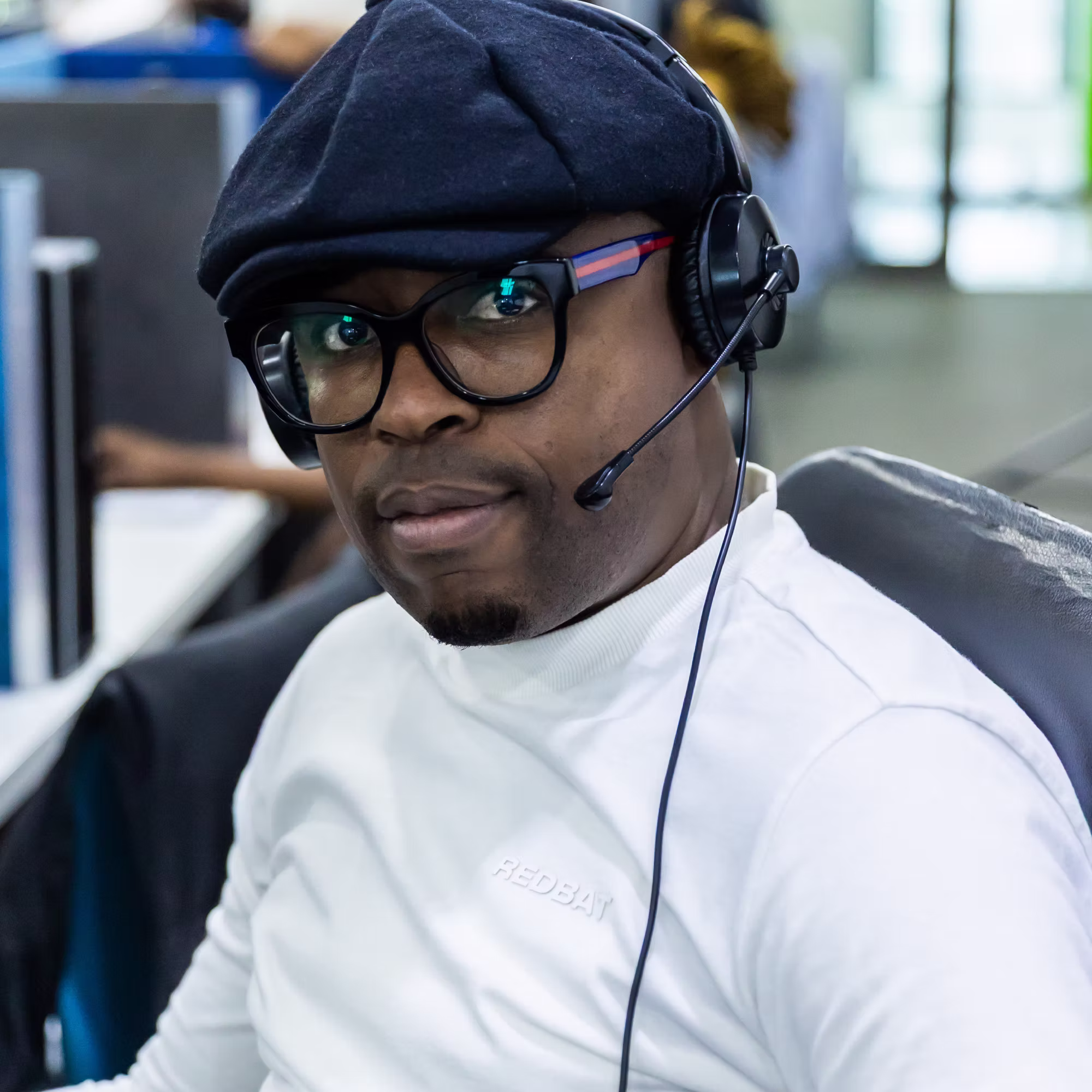 Man wearing glasses, a dark cap, and a headset in an office setting.