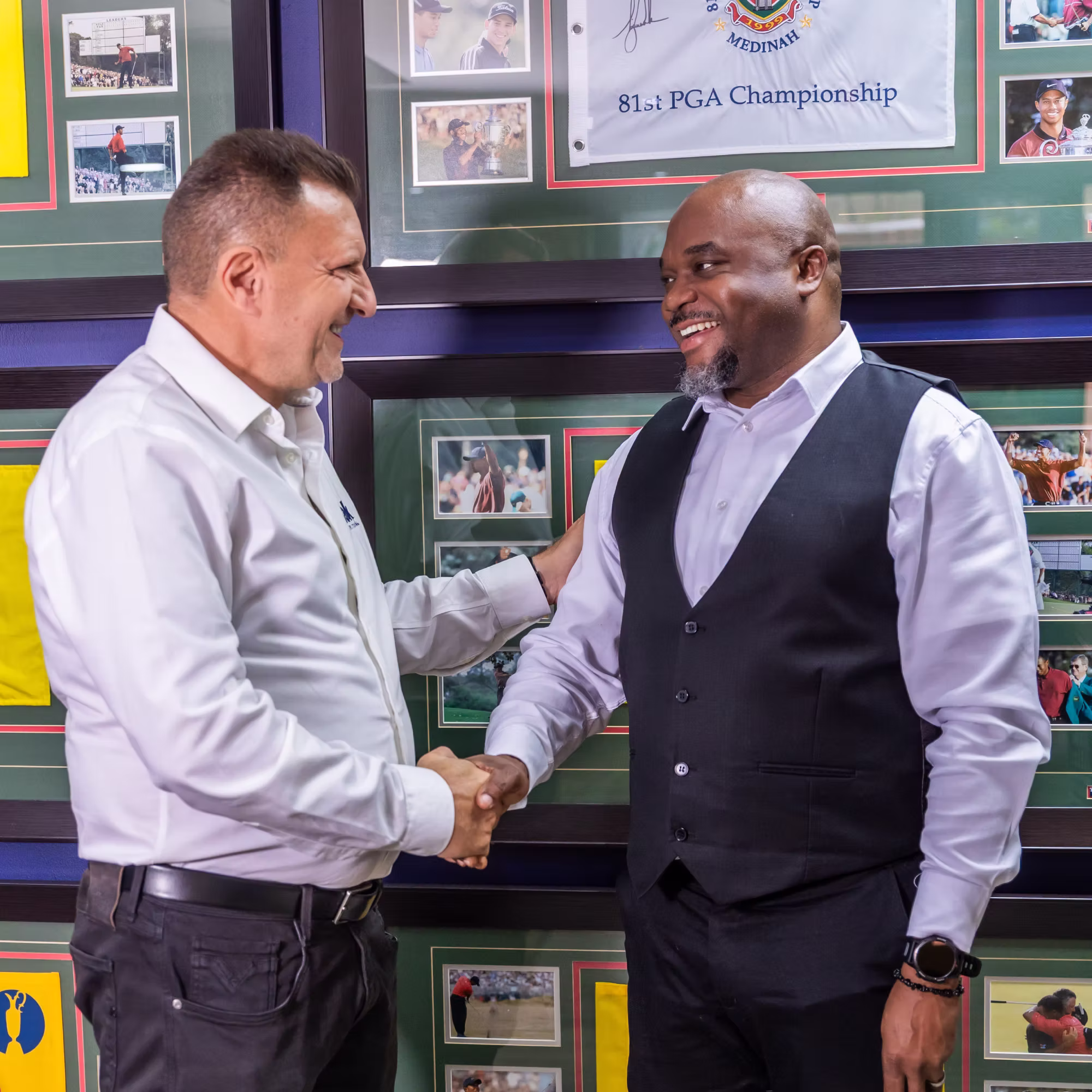 Two men smiling and shaking hands in front of a wall with framed golf photos and memorabilia.