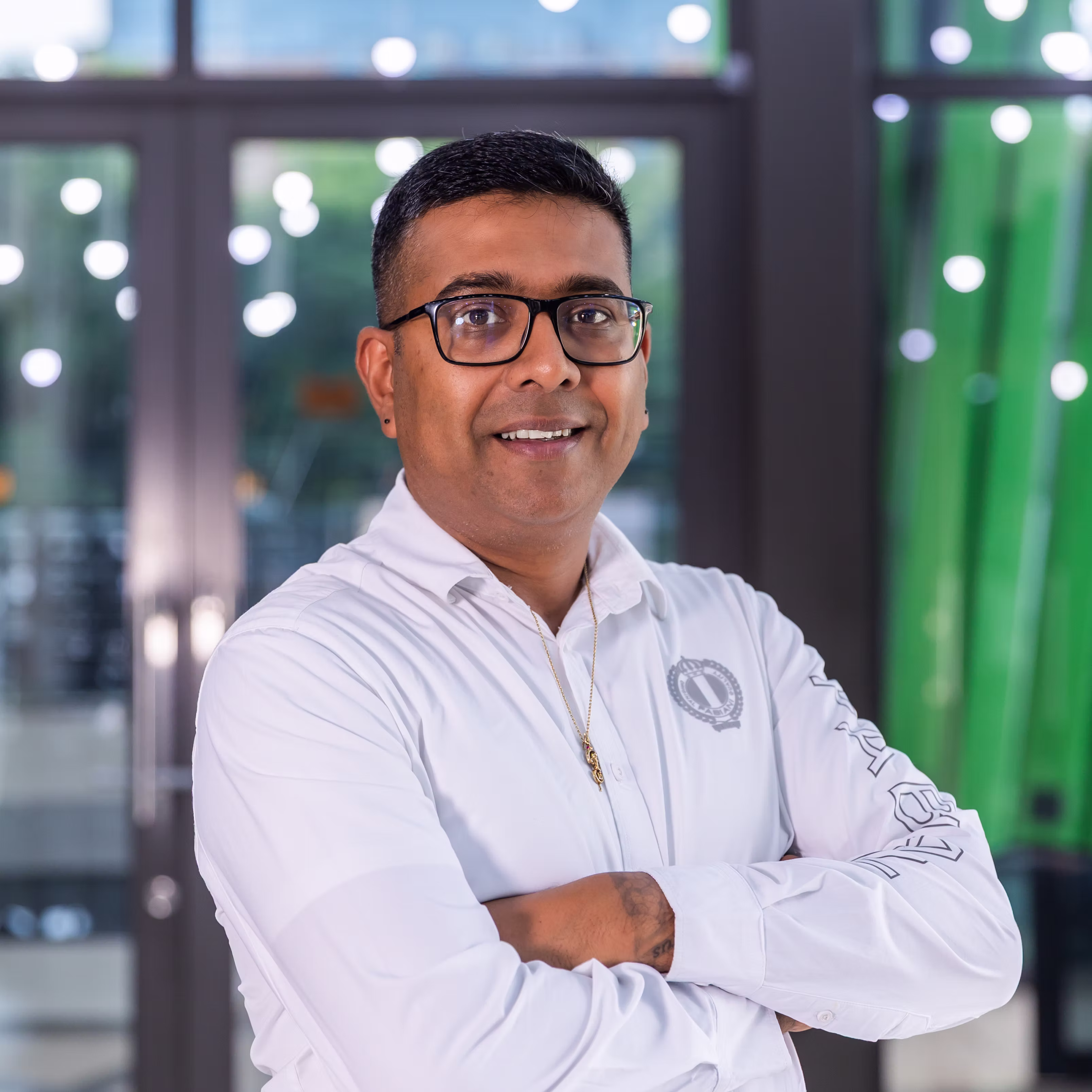 Man with glasses in white shirt crosses arms and smiles before glass wall with round lights.
