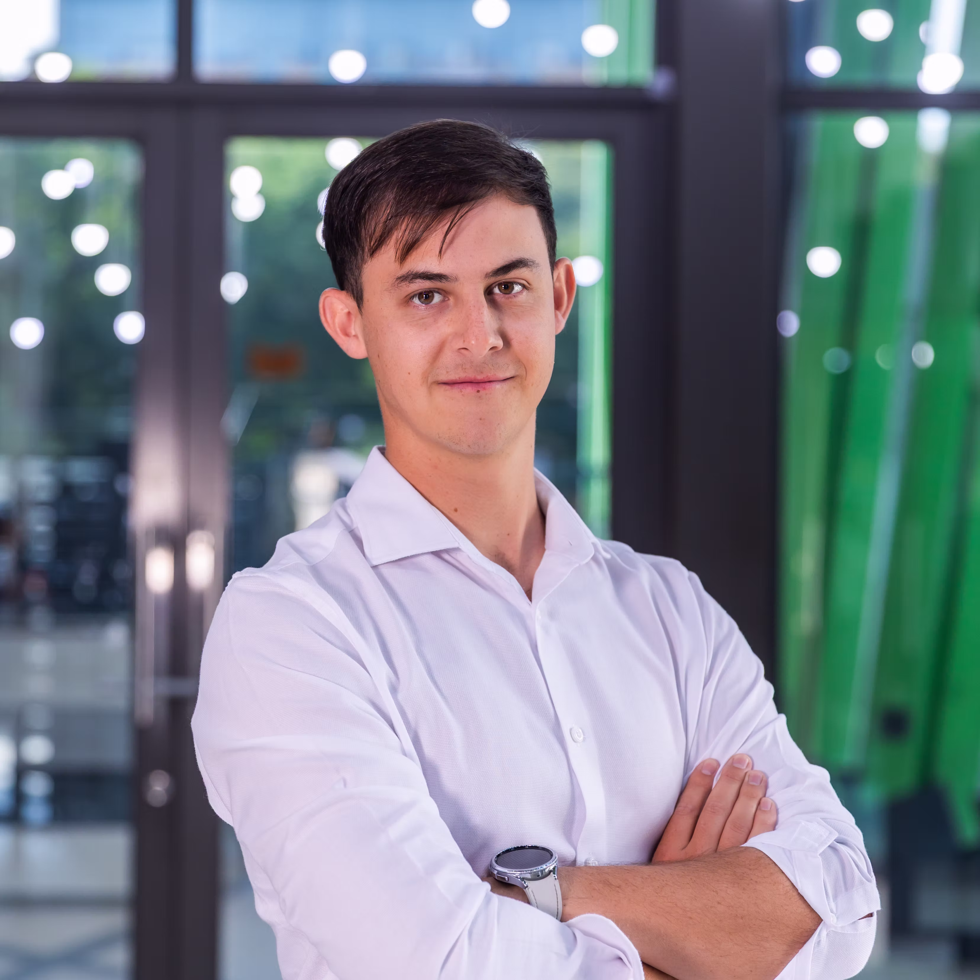 Young man in a white shirt with arms crossed stands in front of glass doors with green decor.