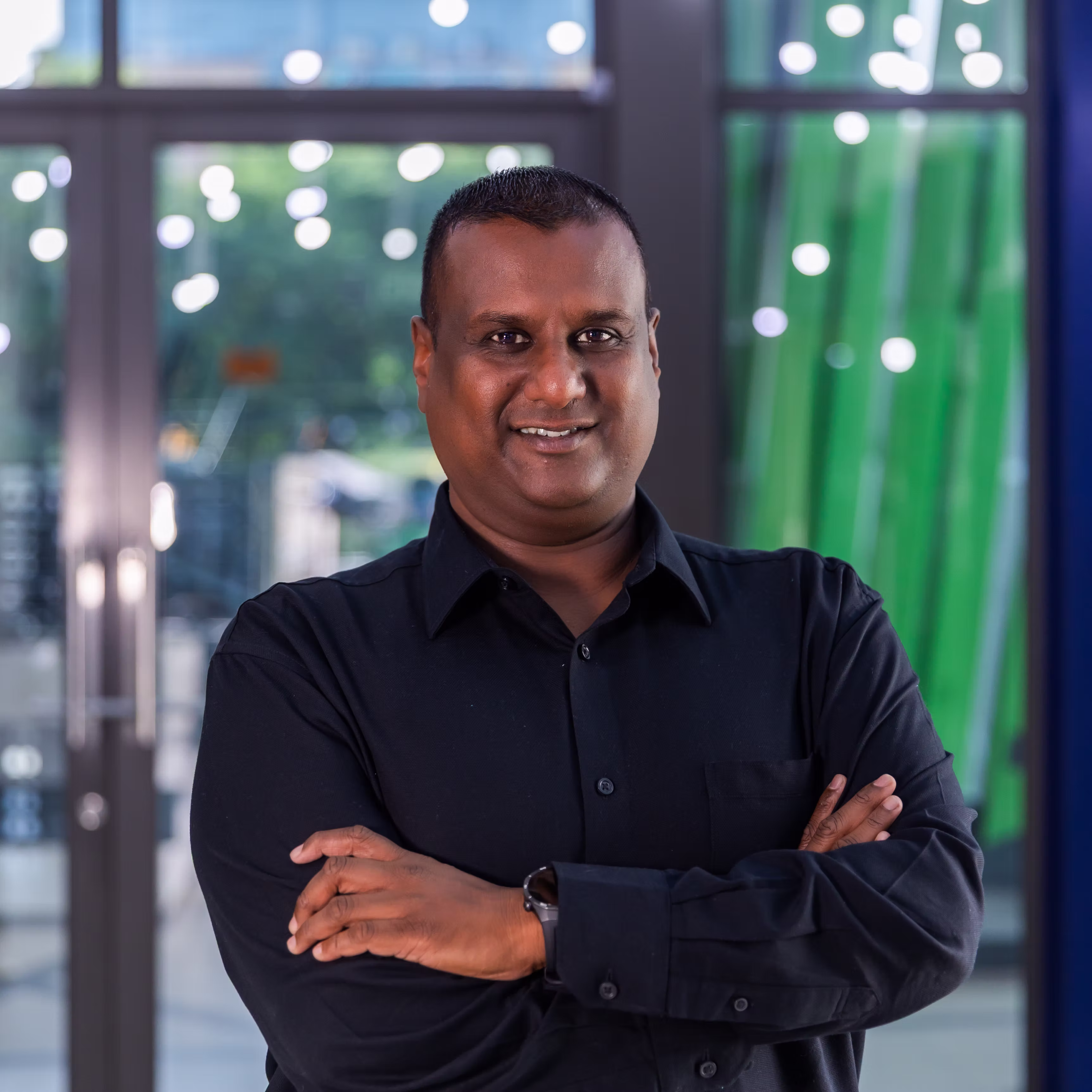 Man in a black shirt with arms crossed smiling inside a modern building with glass doors behind.