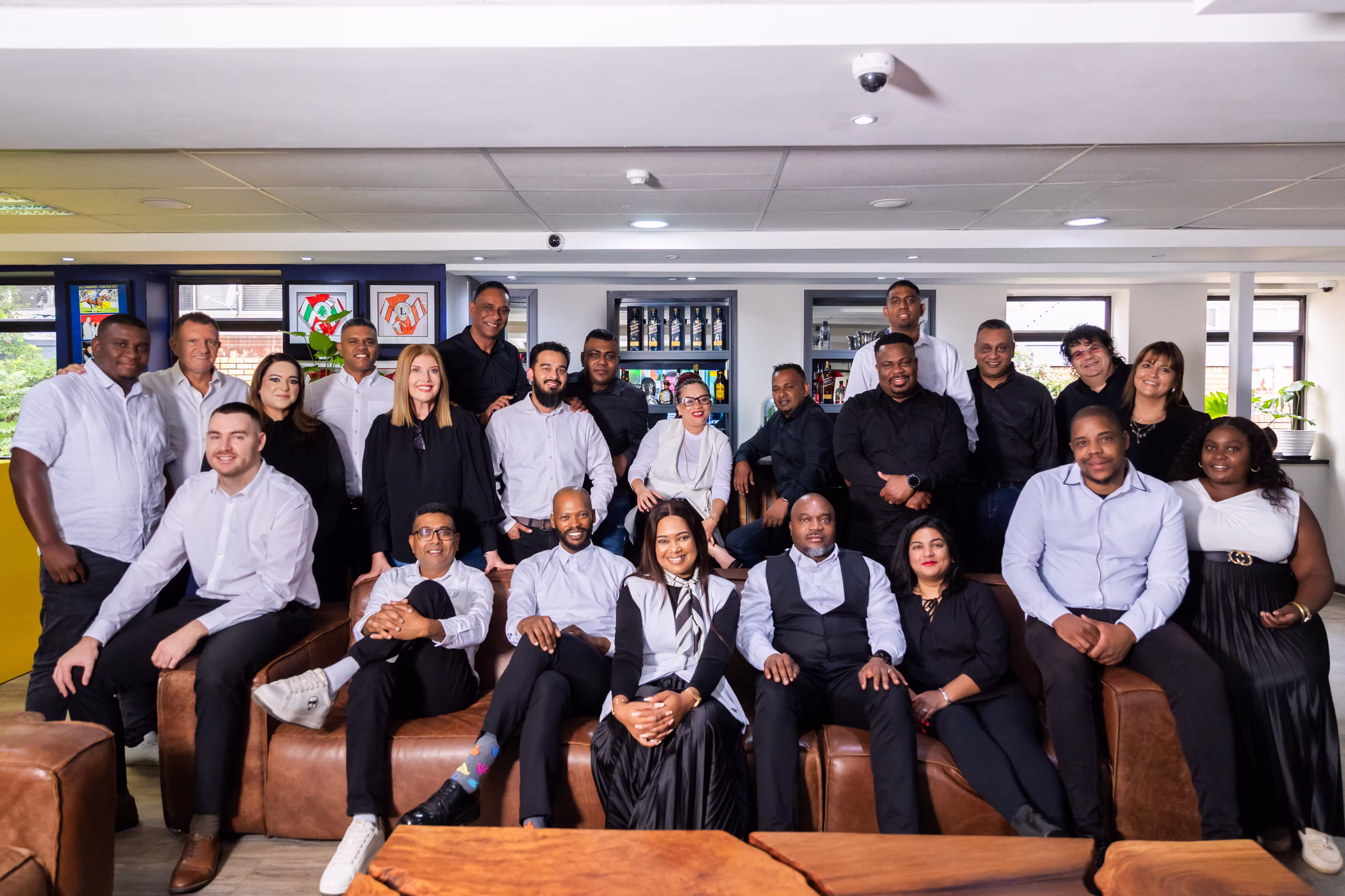 Group of twenty-five diverse people posing for a photo in a modern indoor space with brown leather seating.