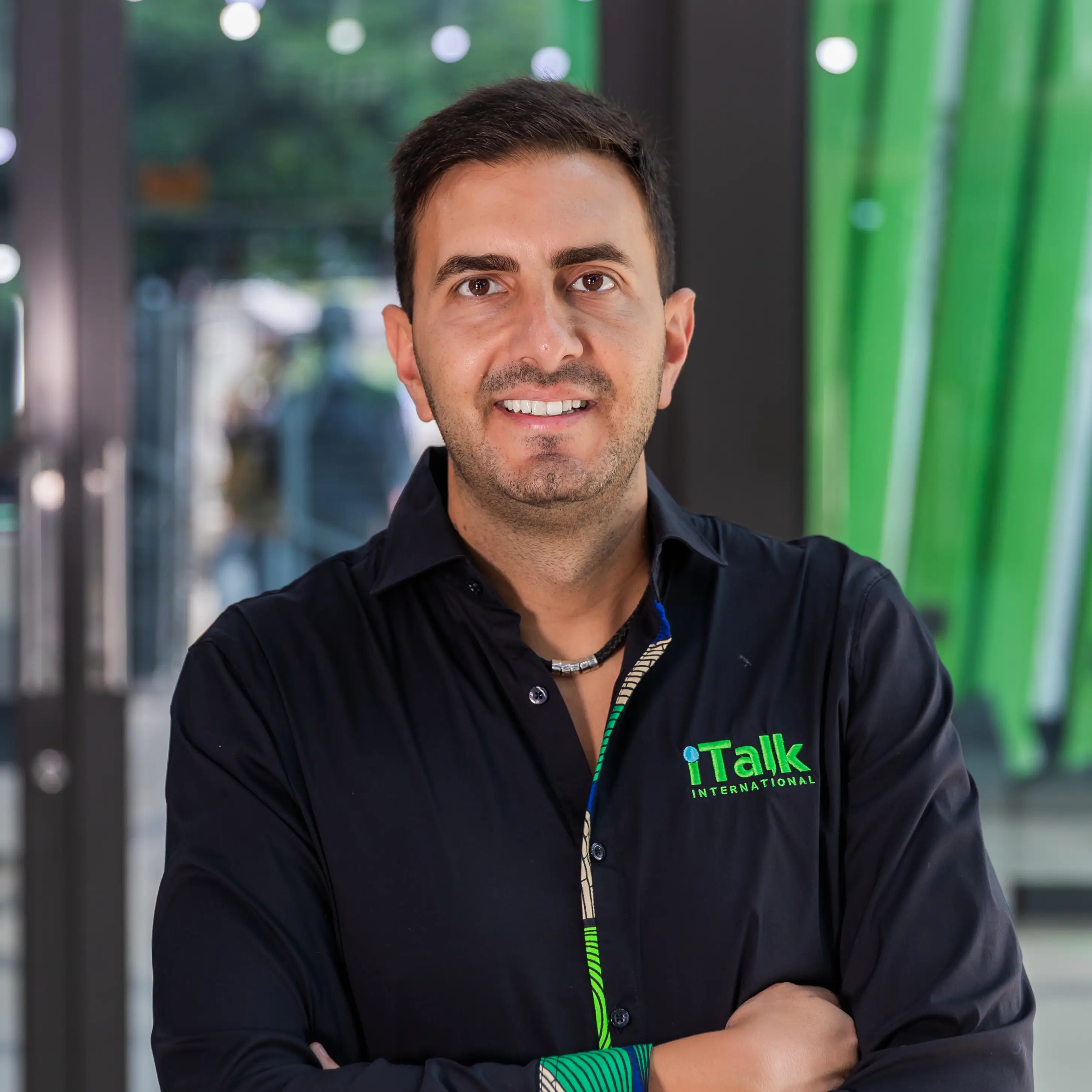 Man with folded arms wearing a black iTalk International shirt in a modern indoor setting.
