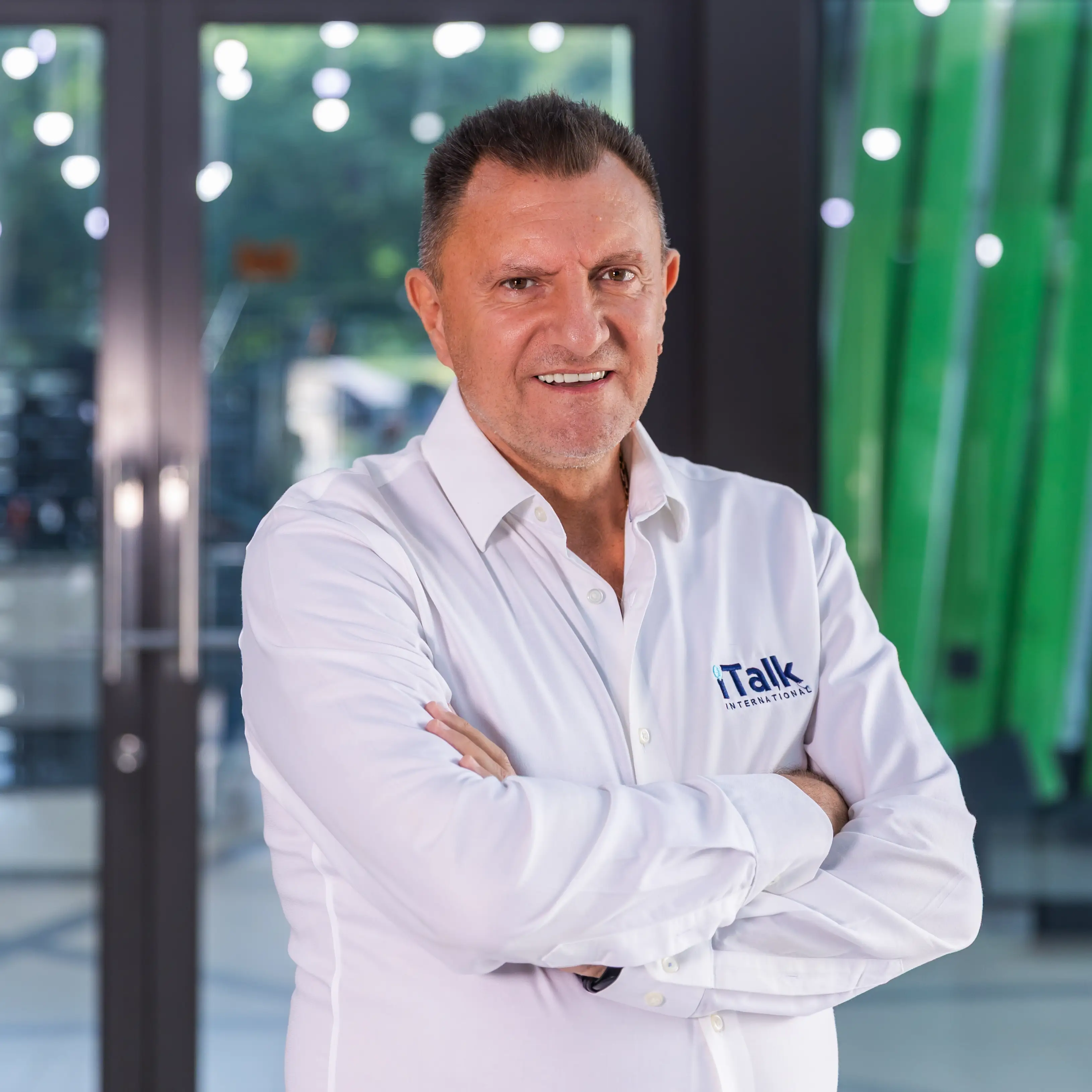Man with short dark hair wearing a white shirt with Talk International logo standing with arms crossed.