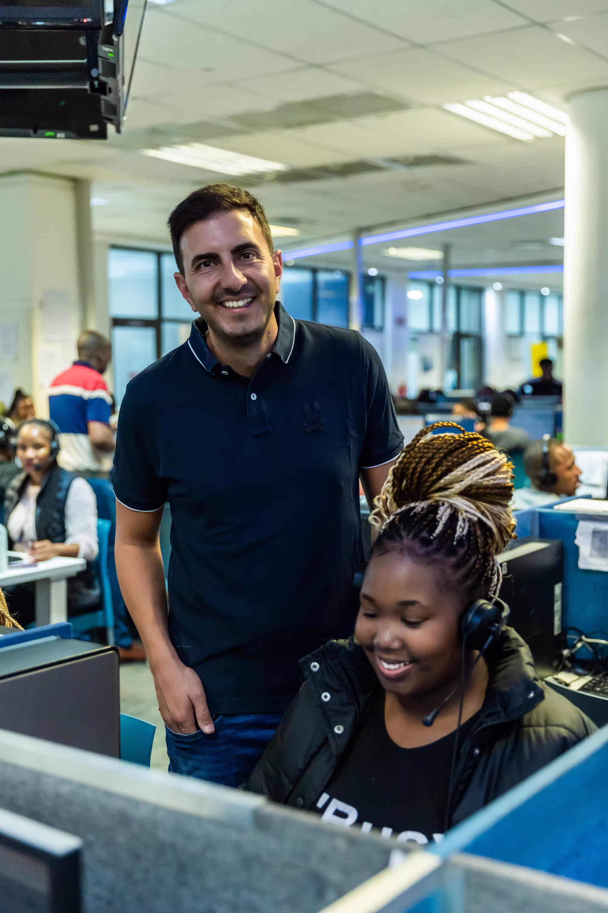 A man stands smiling behind a woman with braided hair wearing a headset in a busy office cubicle.