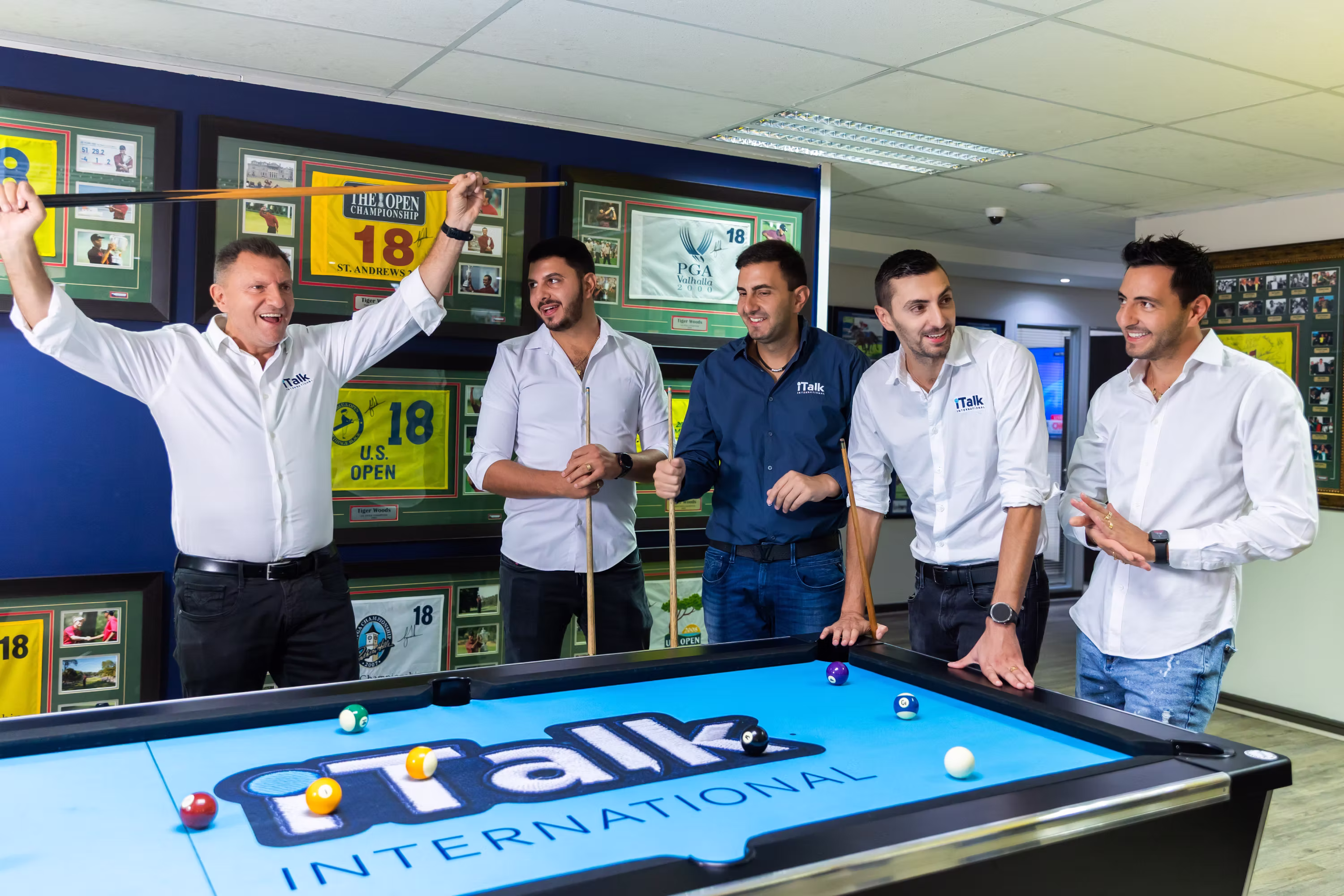 Five men in white and blue shirts stand around a pool table with iTalk International logo, smiling.