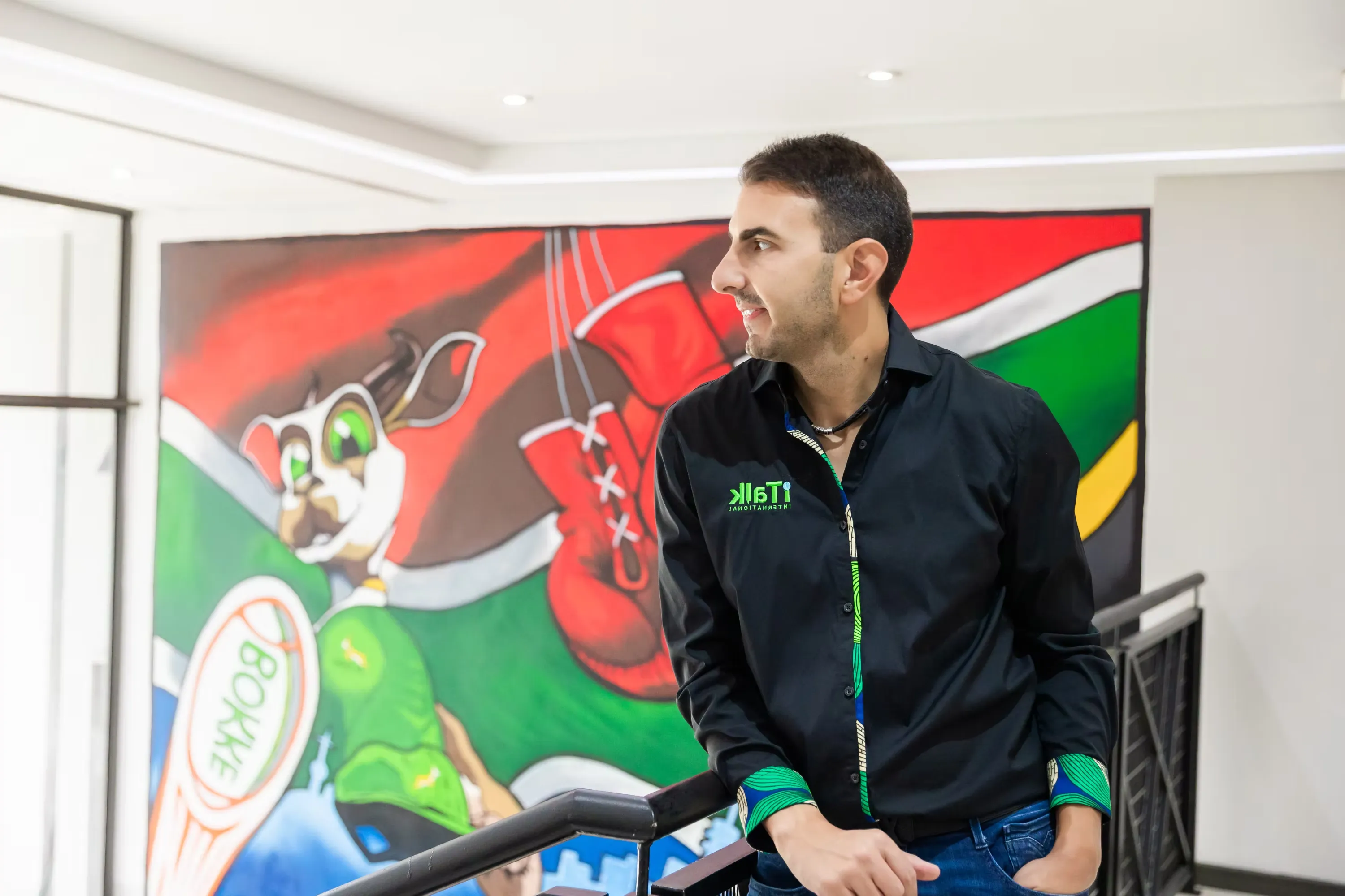 Man in black shirt with iTalk logo leans on railing with colorful cartoon mural in background.