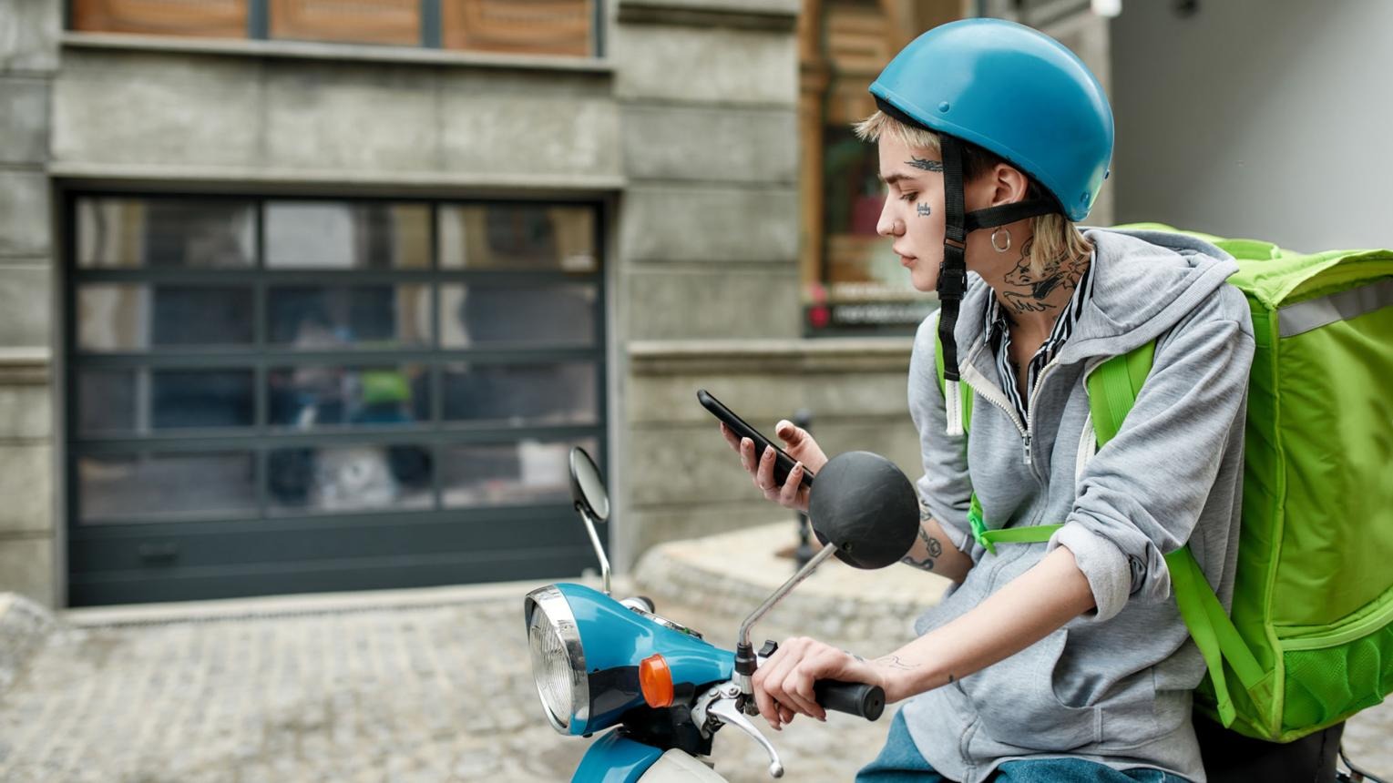 Young woman in helmet with thermo backpack using smartphone while delivering food on a motor scooter