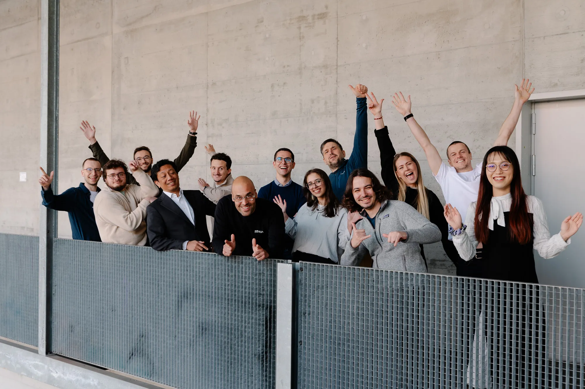 Group of diverse people smiling and raising hands behind a metal fence against a concrete wall.