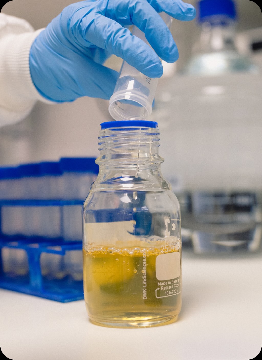 Gloved hand pouring liquid from a small clear container into a large laboratory bottle with yellow liquid inside.