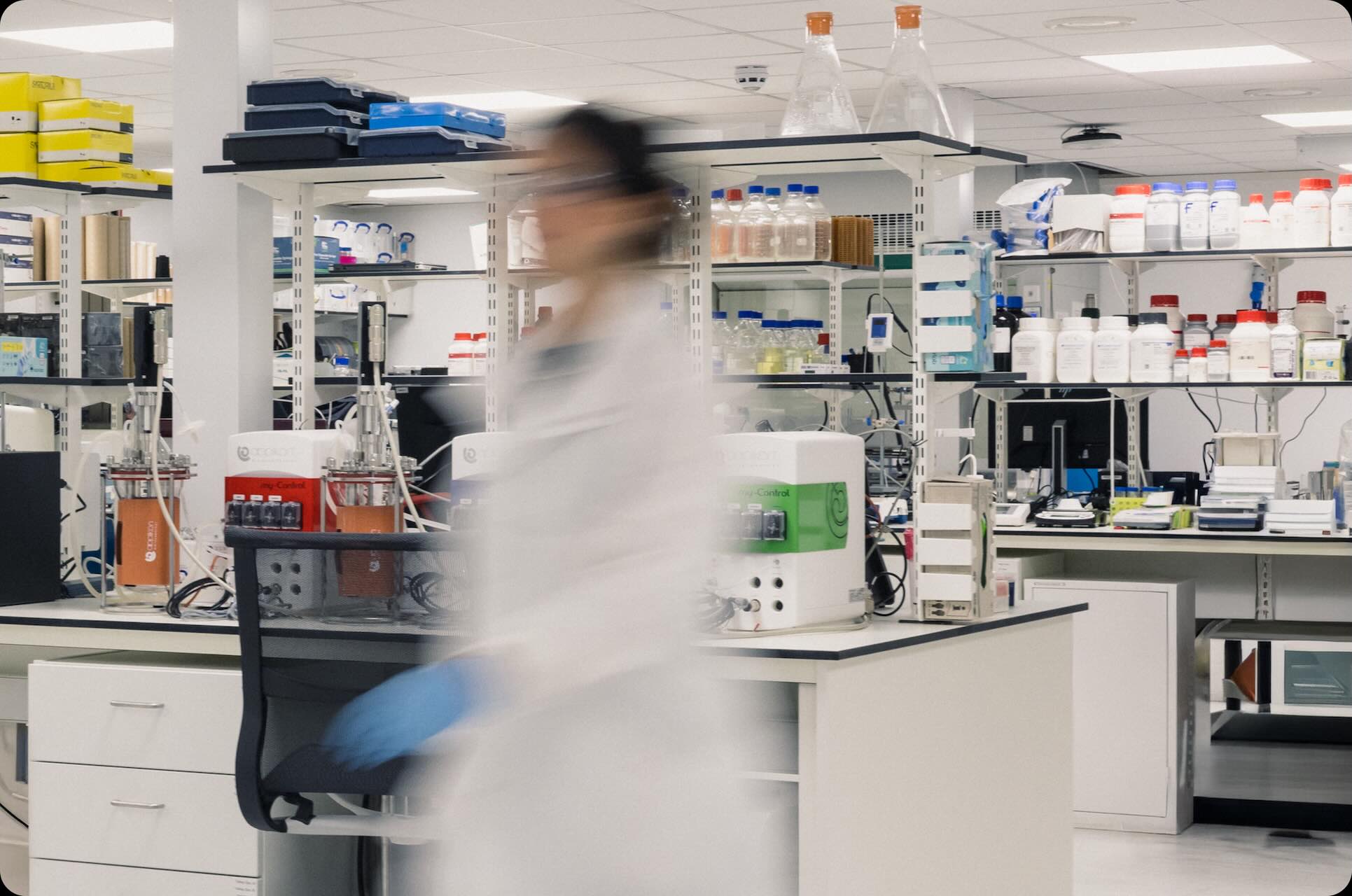 Blurred scientist walking through a modern laboratory filled with equipment, bottles, and supplies.