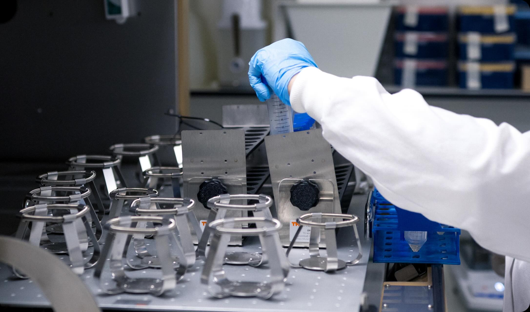 Laboratory technician wearing blue gloves placing a graduated plastic tube into a metal rack on a lab bench.