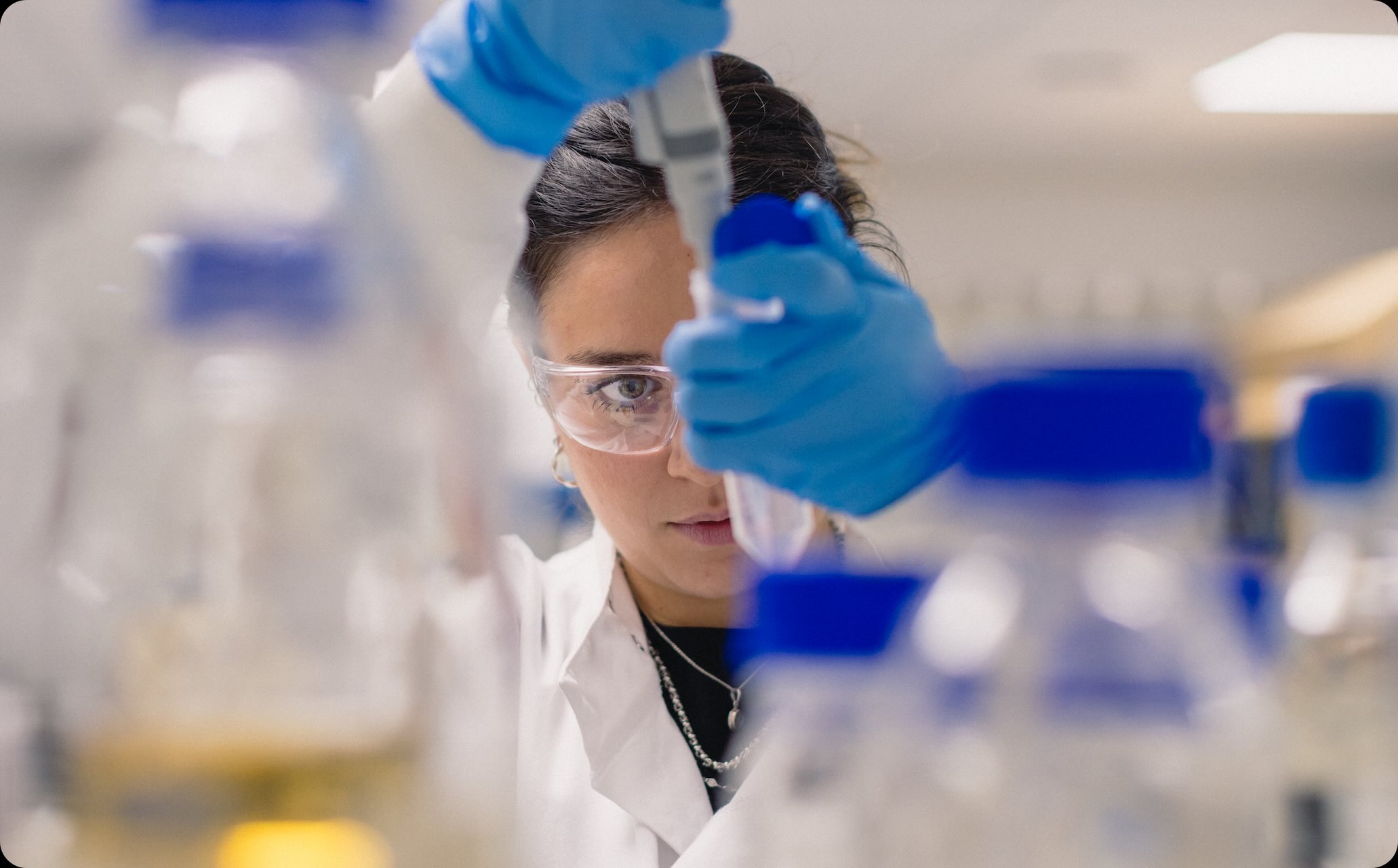Scientist wearing safety glasses and blue gloves using a pipette in a laboratory surrounded by bottles with blue caps.